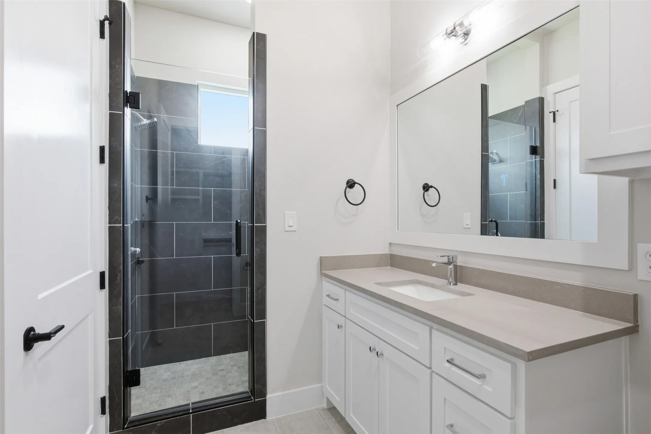Contemporary bathroom featuring a glass-enclosed shower with dark tile surround, a white vanity with a light-toned countertop, and a large framed mirror with overhead lighting