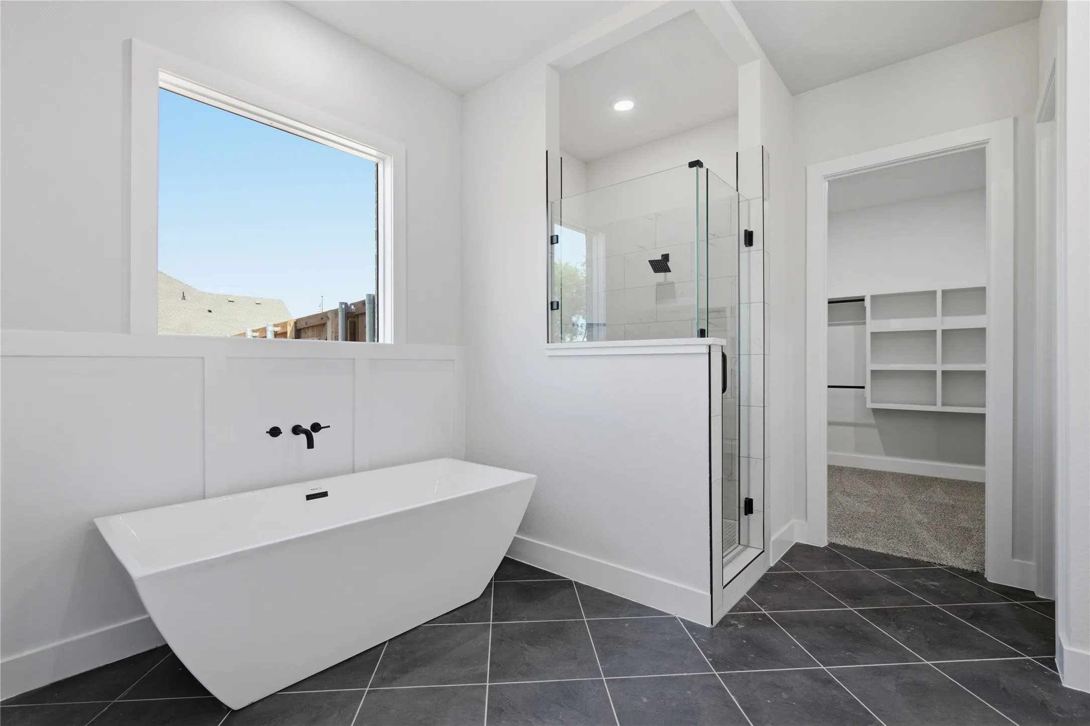 Freestanding soaking tub with wall-mounted matte black faucet, glass-enclosed shower, dark tile flooring, and built-in closet shelving
