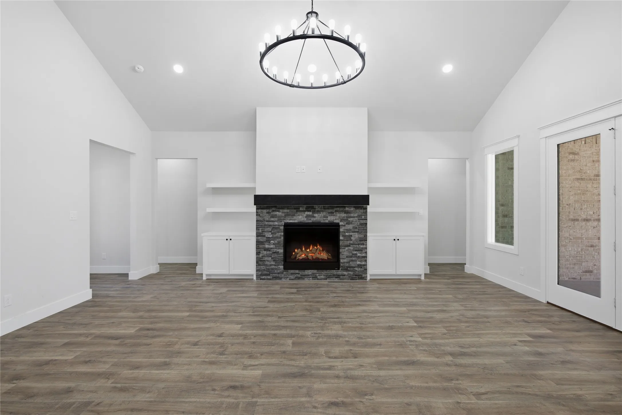 Vaulted ceiling with an architectural chandelier, wood-finish flooring, stone-clad fireplace, built-in shelving, and white French doors