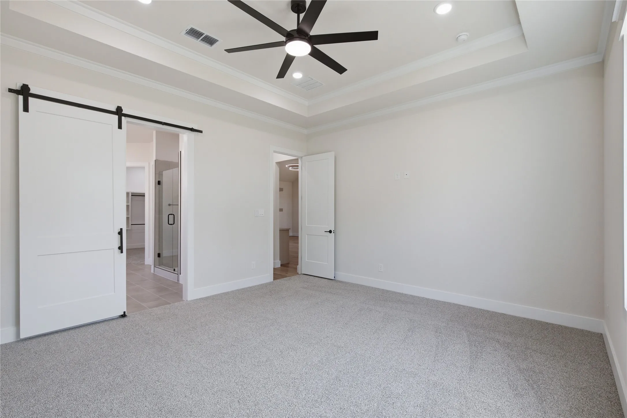 Tray ceiling with recessed lighting and a ceiling fan, light-toned carpeting, white barn-style sliding door, white interior door, and a partially visible glass shower enclosure