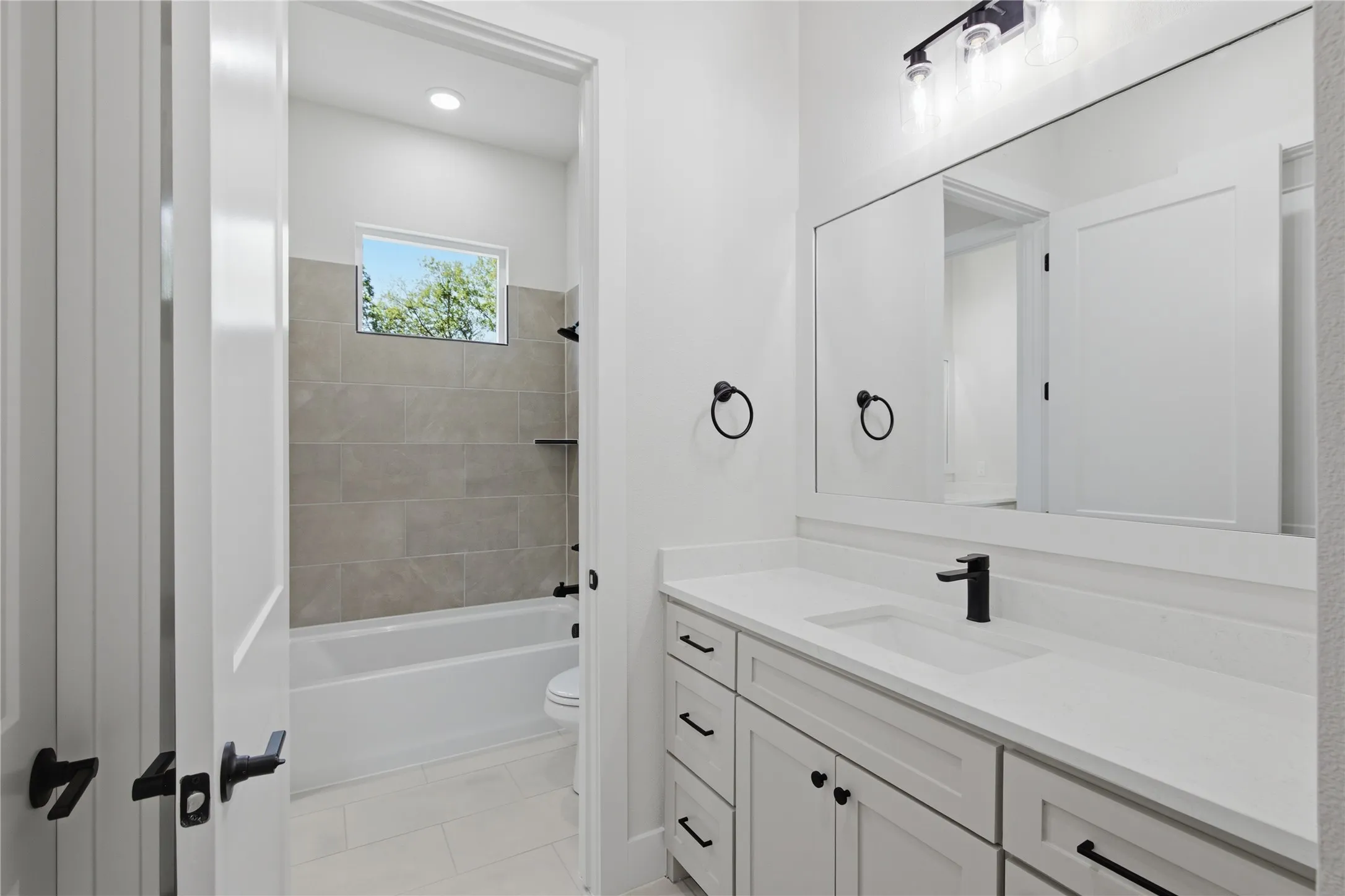 Bathroom featuring a white vanity with integrated sink, matte black fixtures, an expansive wall mirror, a bathtub with a tiled surround, and recessed lighting