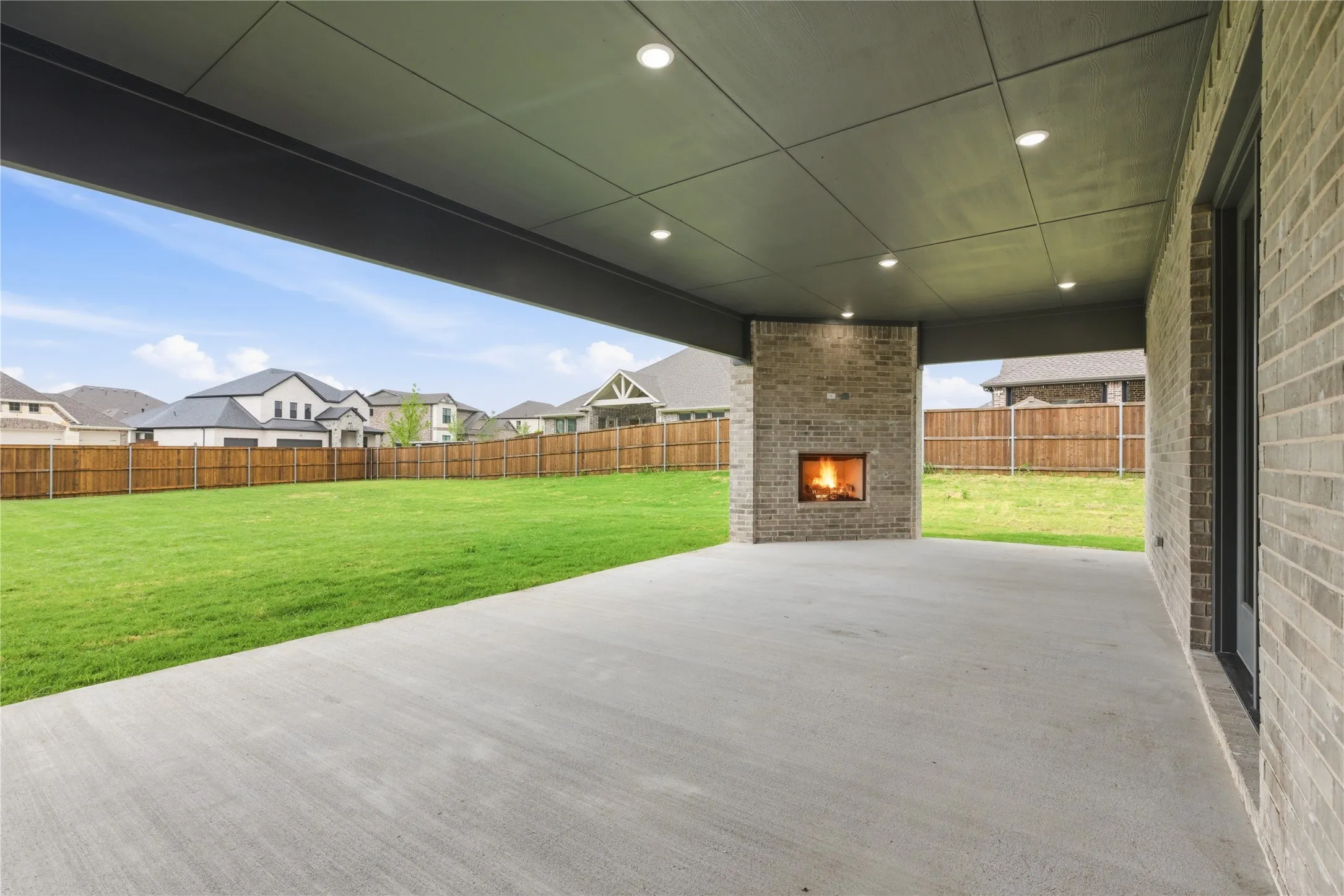 Covered outdoor patio featuring a brick-clad fireplace, recessed lighting, concrete flooring, and an expansive lawn