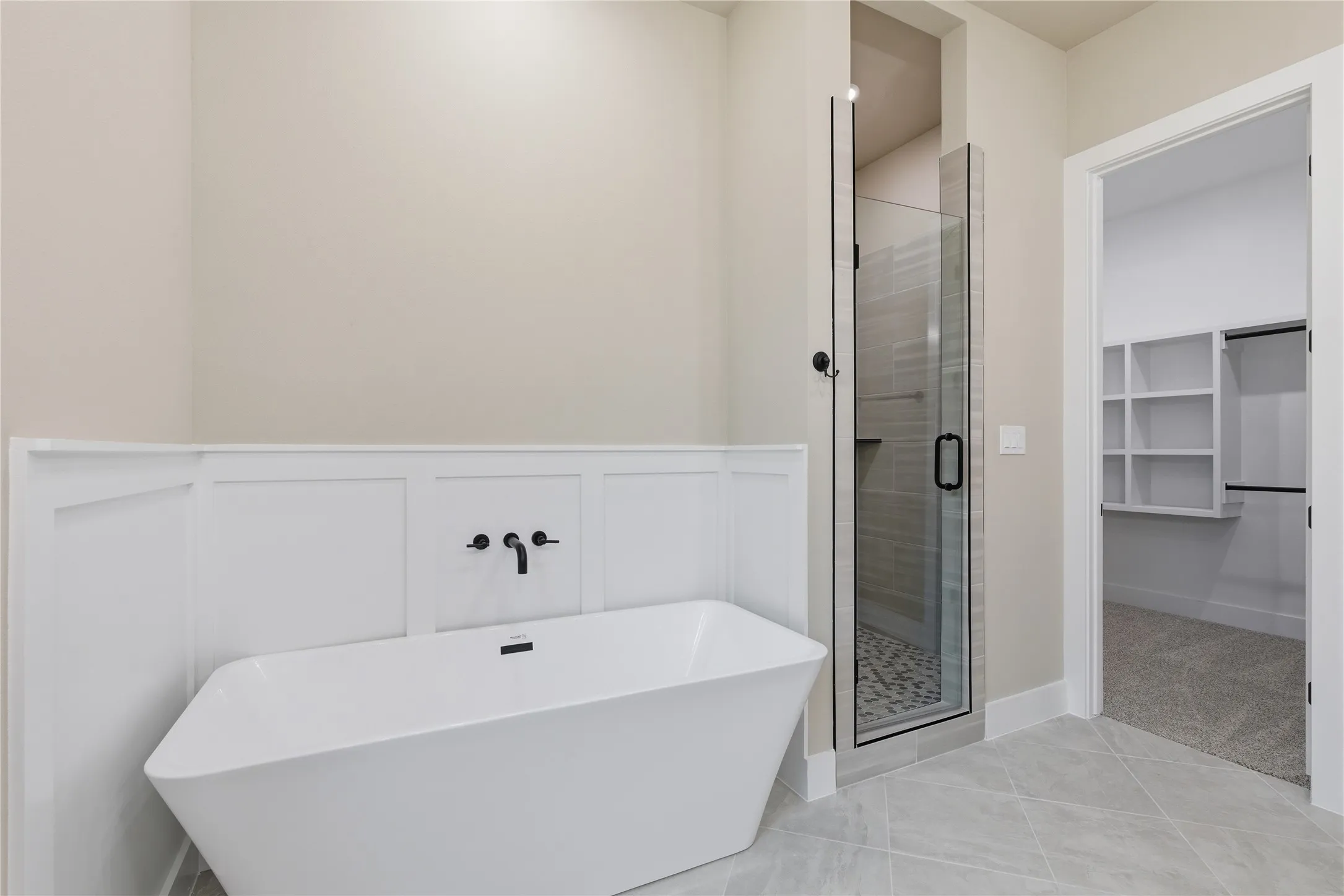 Freestanding soaking tub with wall-mounted matte black fixtures, glass-enclosed shower with tiled floor, and an adjacent walk-in closet featuring built-in shelving