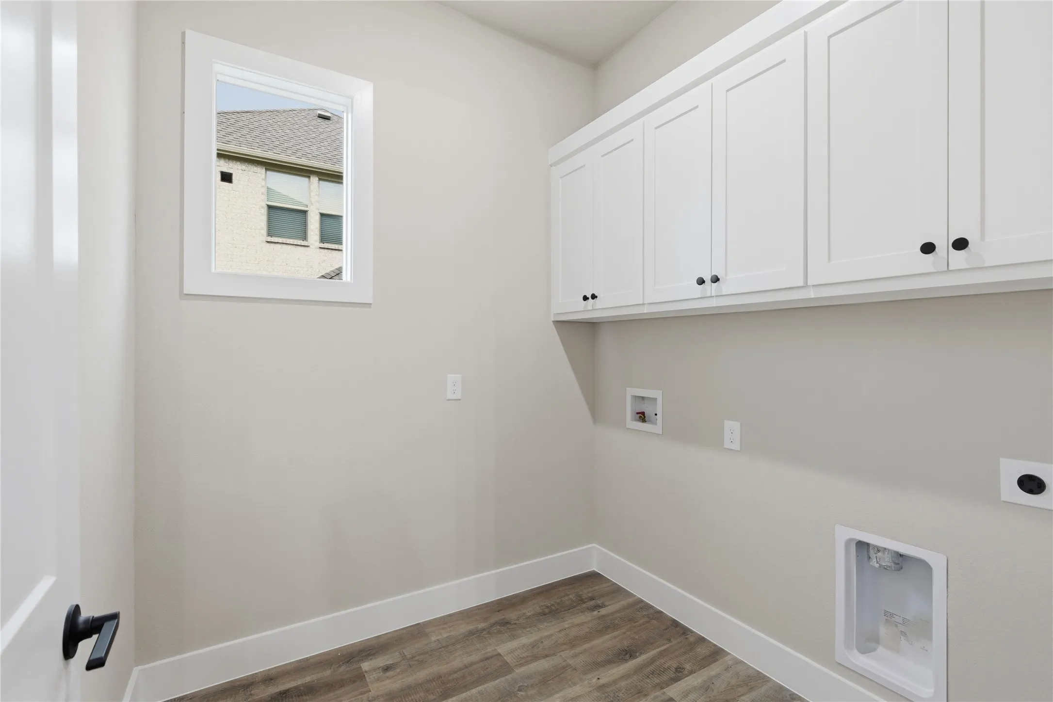 Utility room featuring white shaker-style cabinetry with black hardware, wood-finish flooring, and a single window with white trim