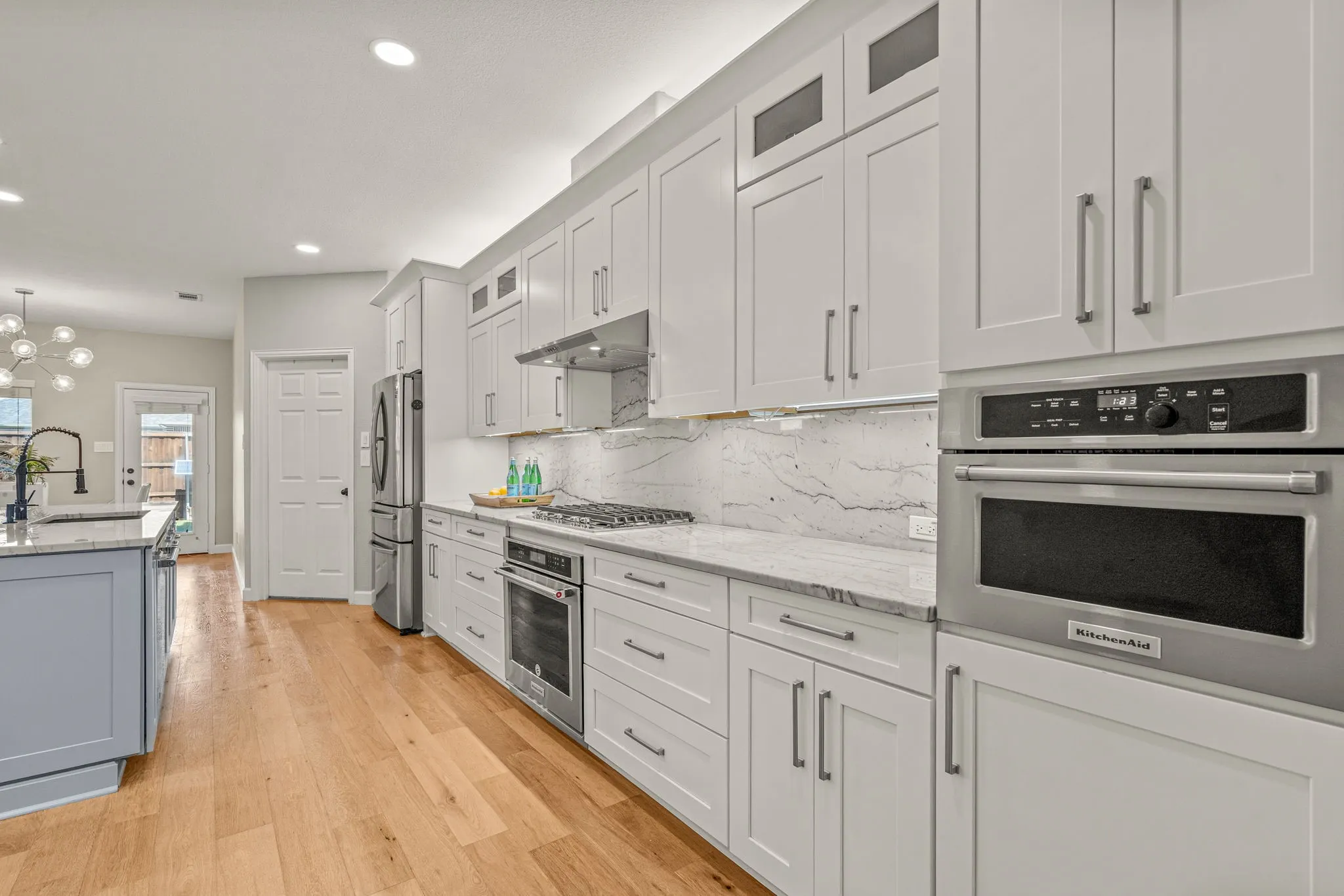 Kitchen with glass fronted cabinets, stainless steel appliances, light stone countertops, light wood-style floors, and white cabinetry