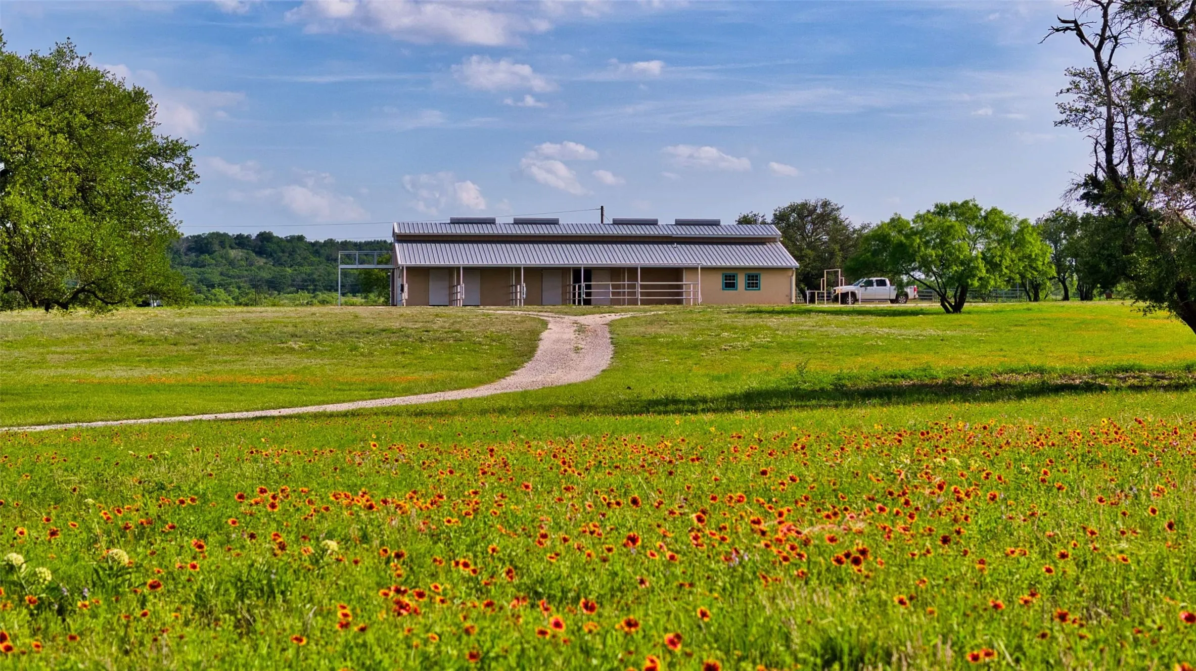 Expansive grassy field featuring a meandering dirt path leading to a single-story building with a metal roof and light-colored siding