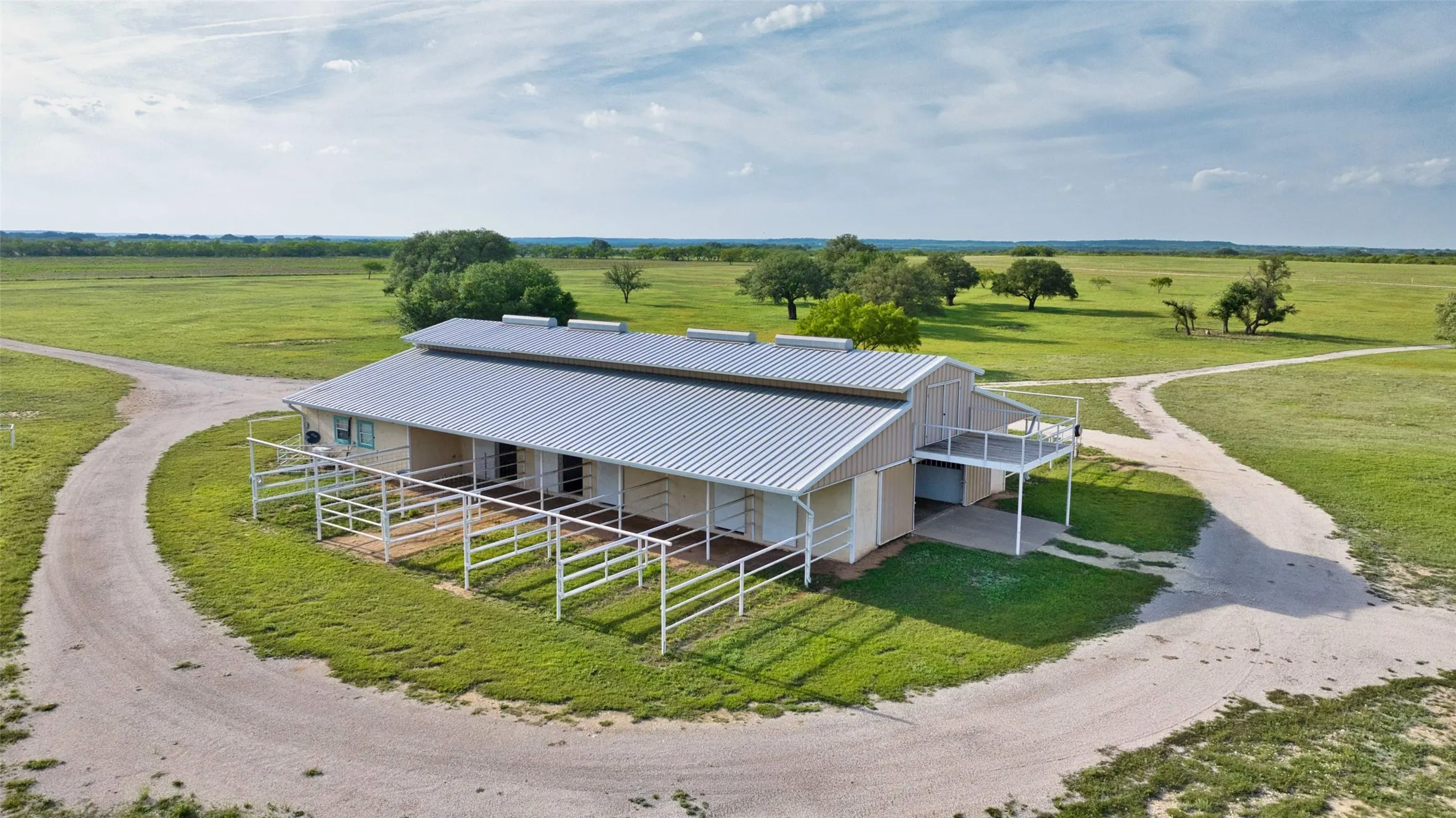 Expansive barn structure featuring a metal roof, multiple stalls with white fencing, a second-story balcony, and a gravel driveway, all surrounded by open green fields