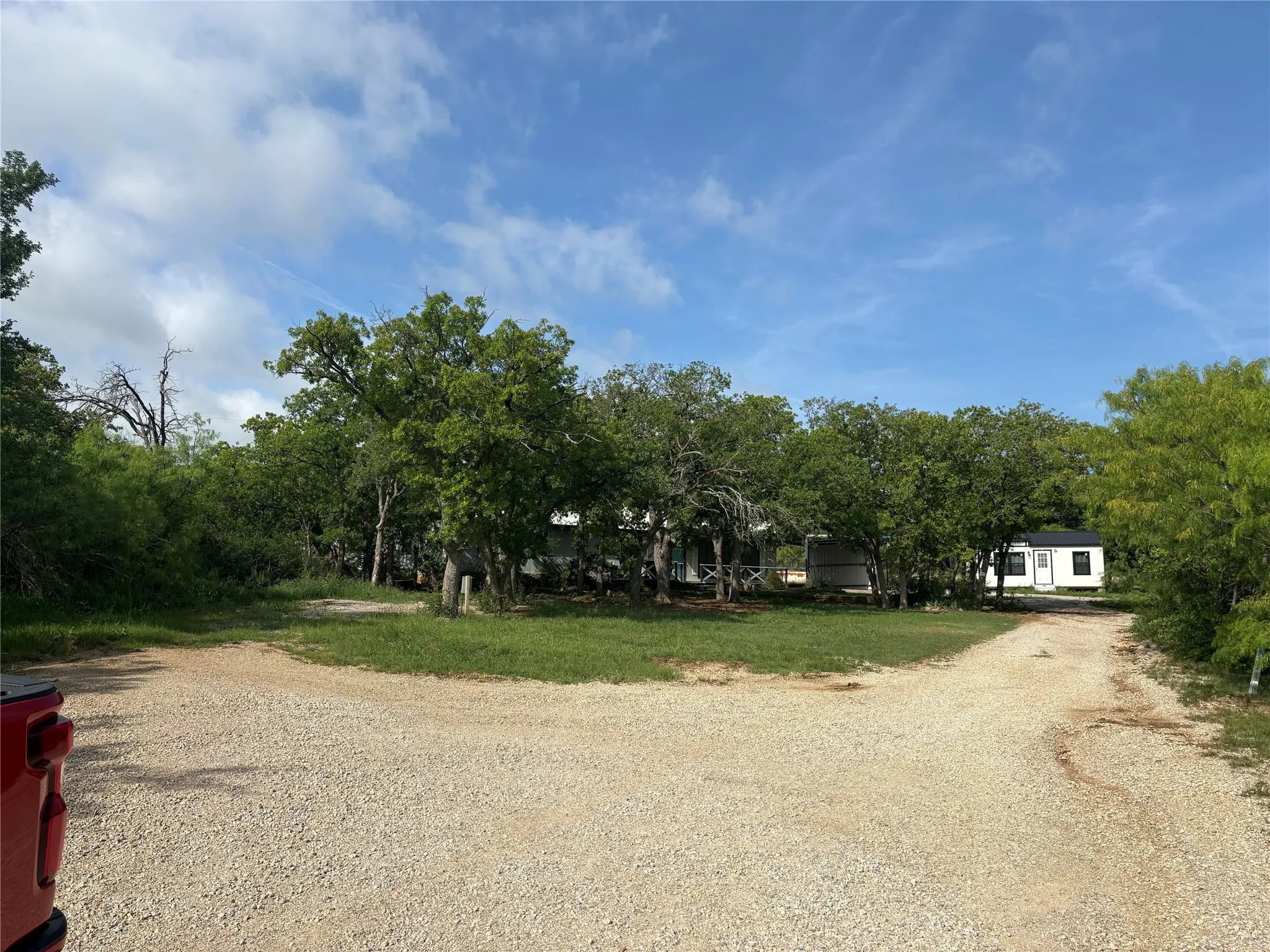 The property features a gravel driveway, a grassy area, and mature trees, with various structures visible through the foliage