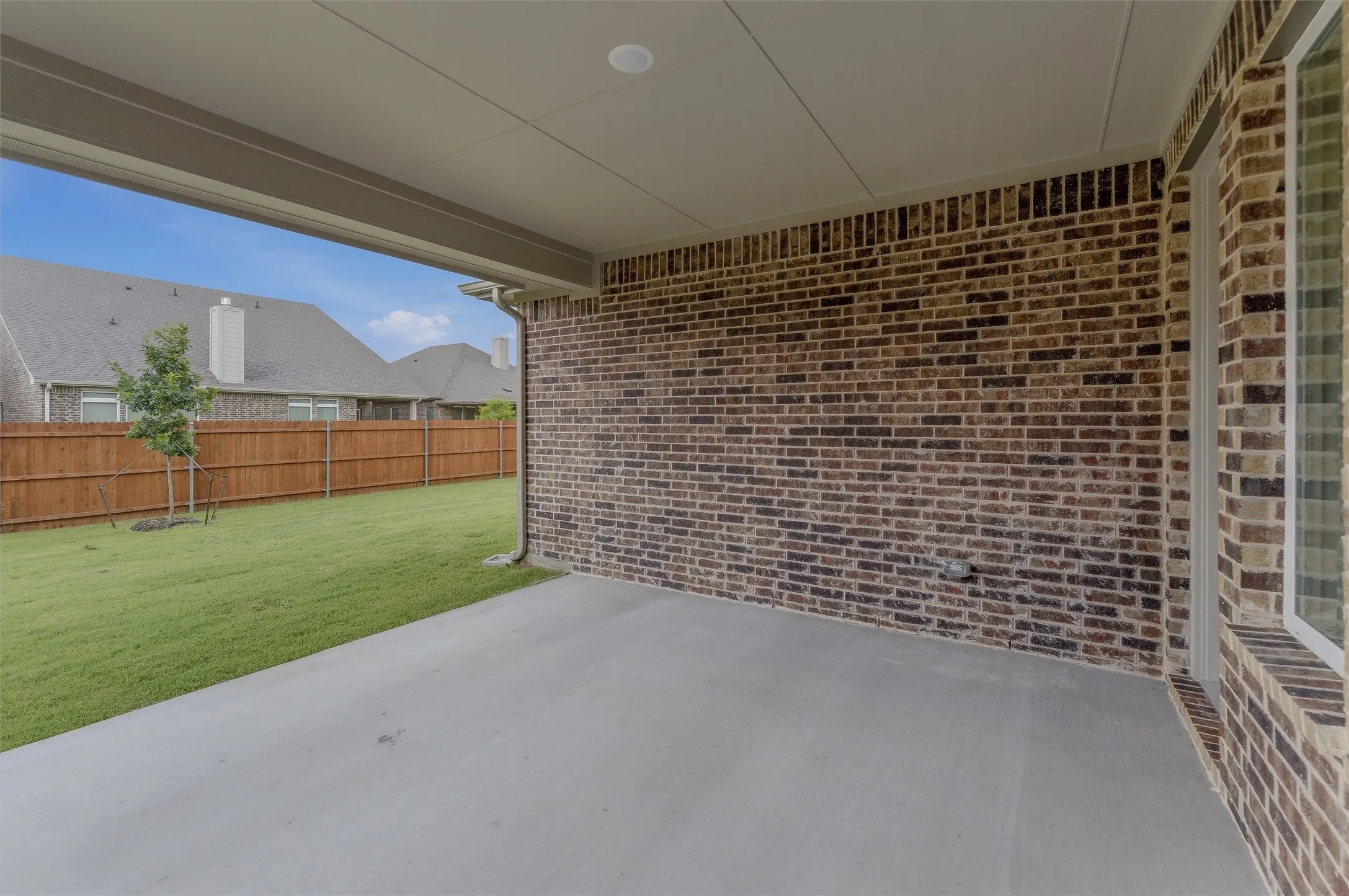 Covered concrete patio featuring a brick exterior wall, recessed lighting, and a view of the fenced backyard with green lawn