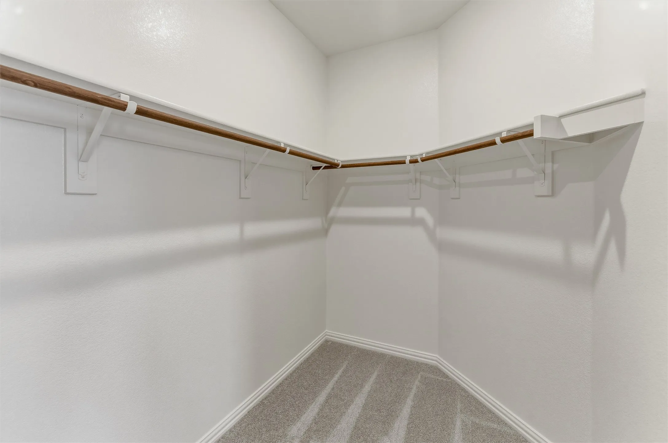 Walk-in closet featuring light gray carpet flooring, white painted walls, and a corner shelving system with integrated wood-finish hanging rods