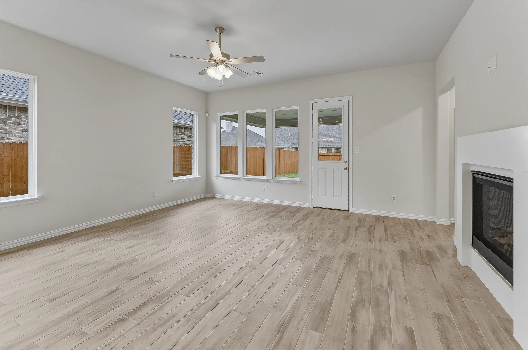 Spacious room featuring light wood-finish flooring, multiple windows, a ceiling fan with integrated lighting, a glass-paned exterior door, and a built-in fireplace with a white surround