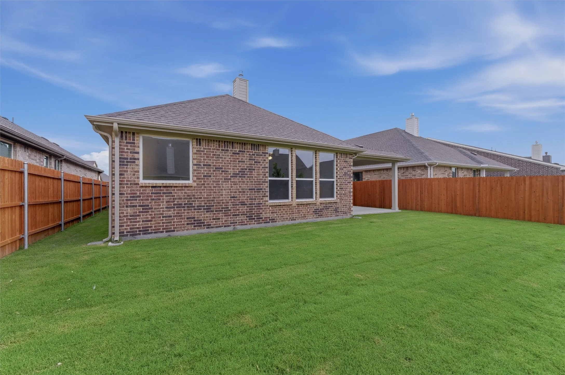 Expansive green lawn with a brown privacy fence, brick exterior, multiple windows, and a covered patio area