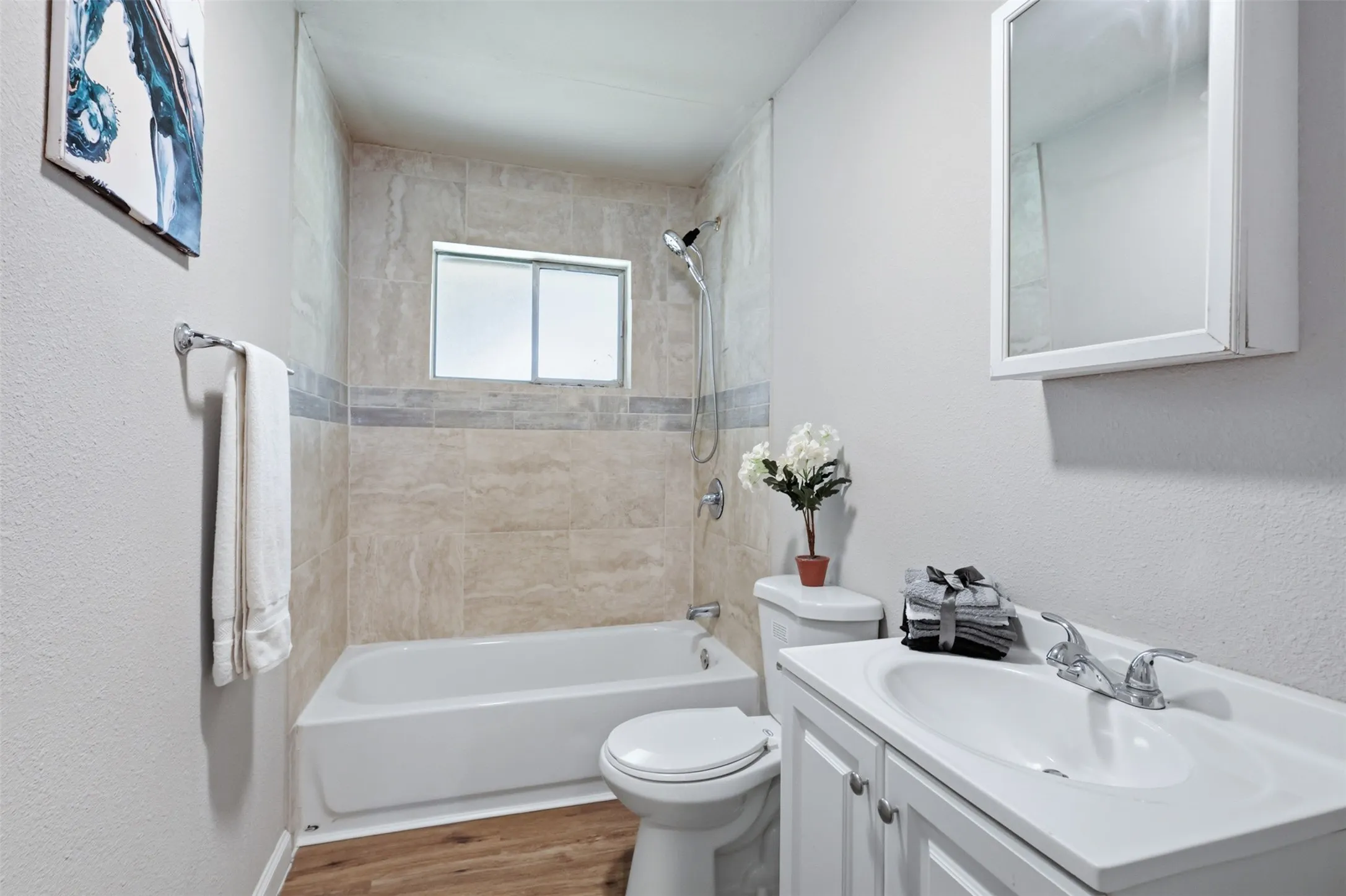 Bathroom featuring a tub/shower combination with natural stone-look tiling and decorative accent band, a white vanity with integrated sink, chrome fixtures, and laminate flooring.