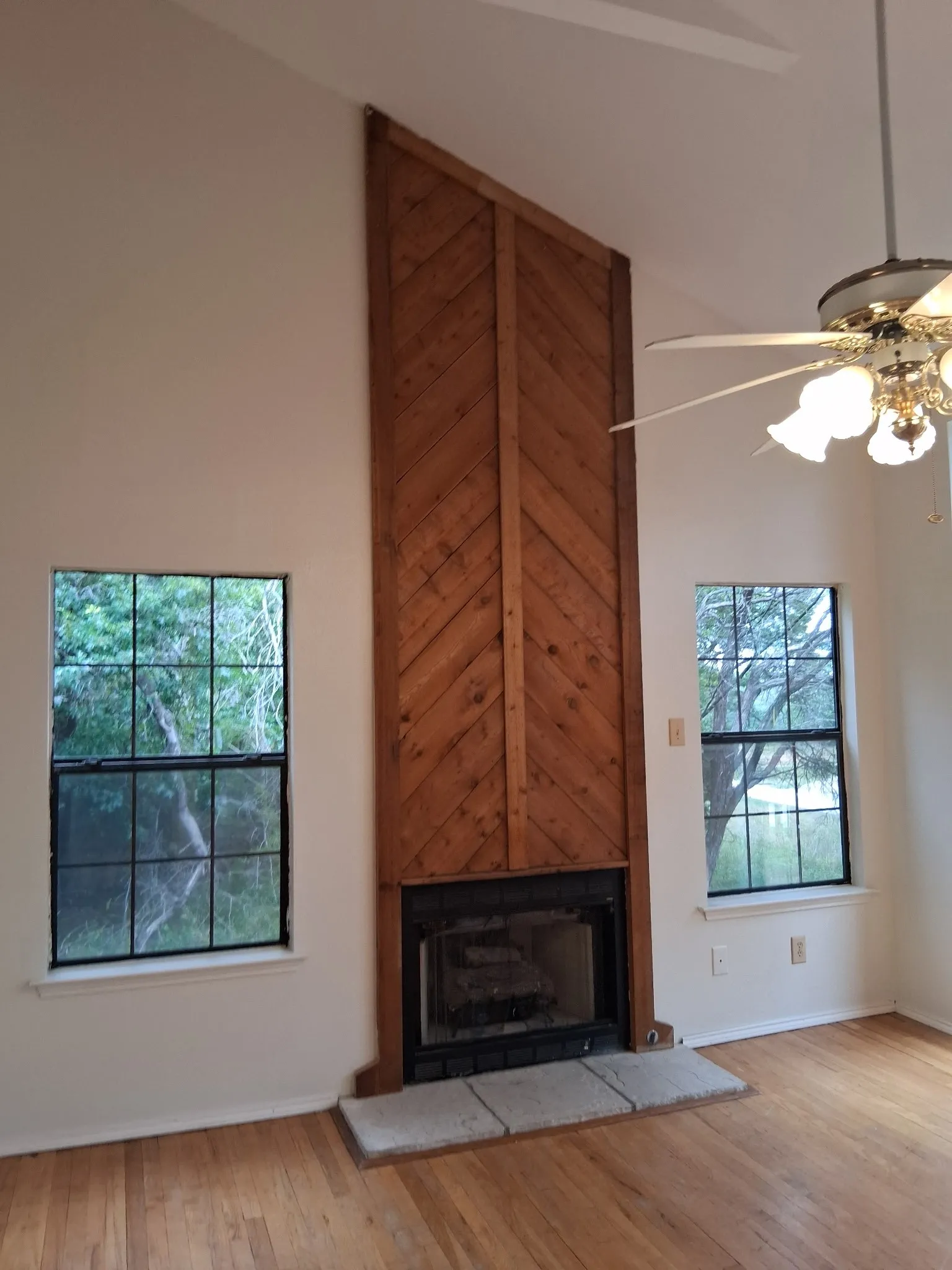 Vaulted ceiling living space featuring a wood-burning fireplace with a wood-paneled surround, two large windows, wood-finish flooring, and a ceiling fan with integrated lighting