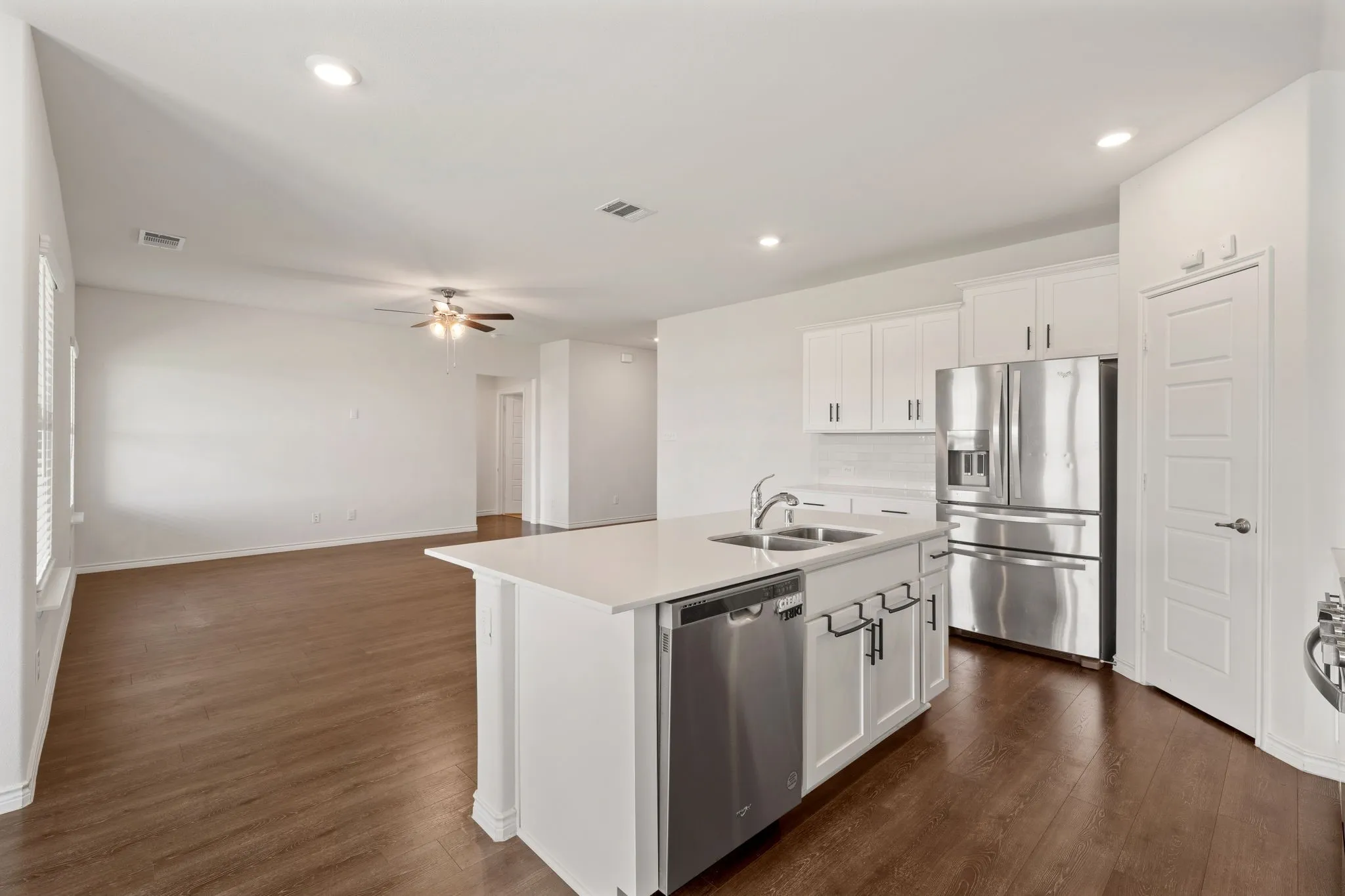 Open-concept living space featuring a central kitchen island with a double basin sink, white cabinetry with dark hardware, stainless steel appliances, and wood-finish flooring