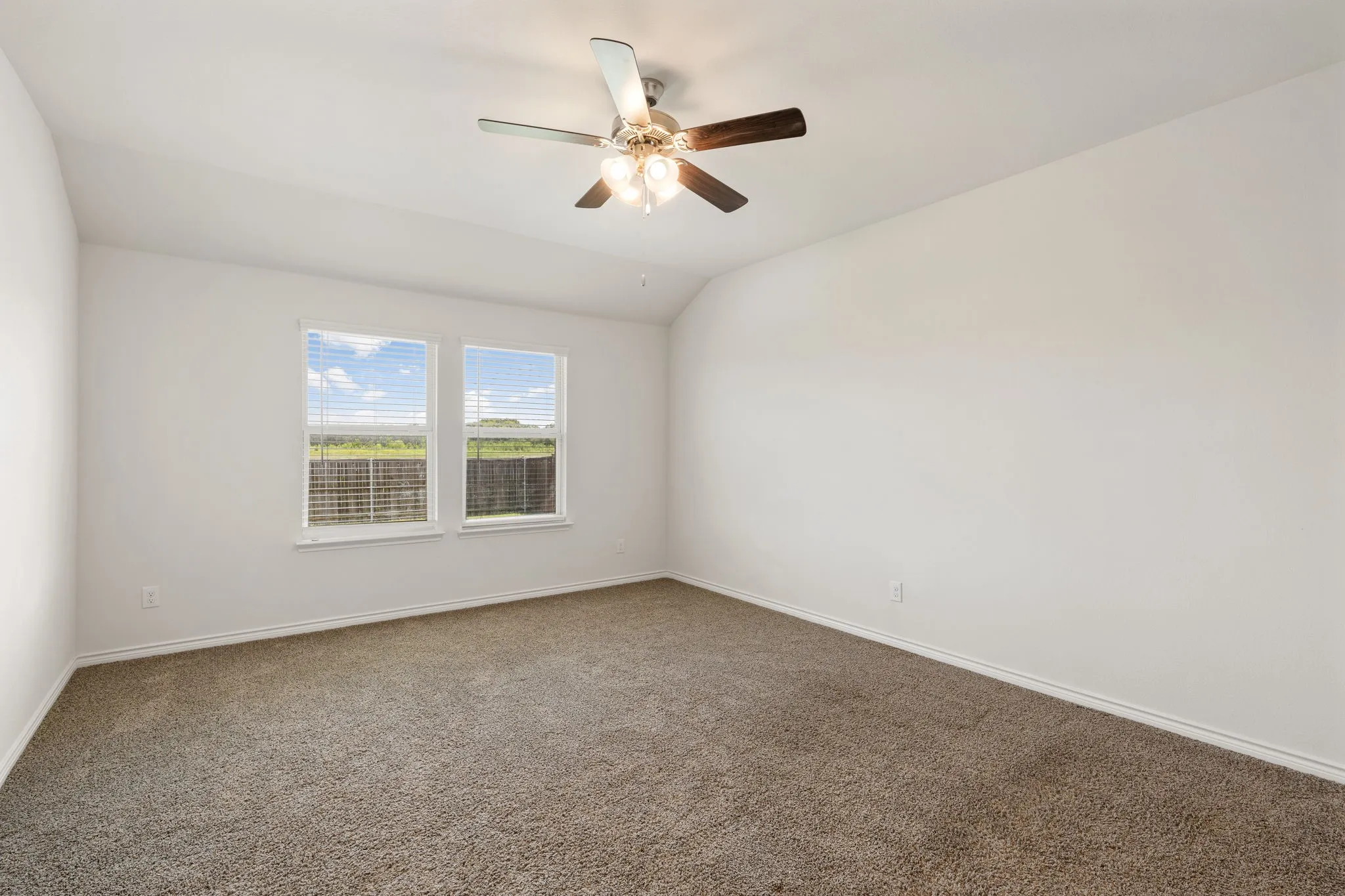 Spacious room featuring neutral carpet flooring, white walls, two double-hung windows with blinds, and a ceiling fan with integrated lighting
