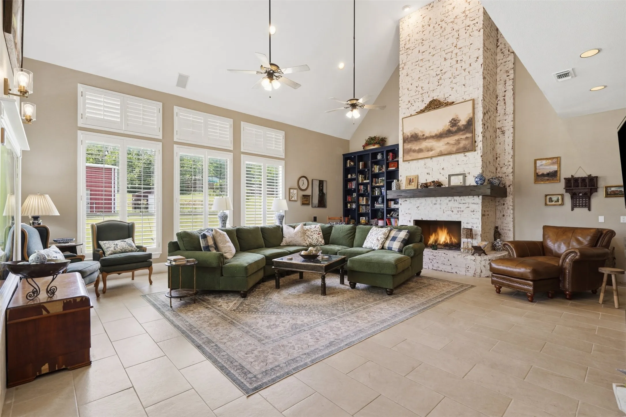 Vaulted living space featuring a white-washed brick fireplace with a substantial wood mantel, tall windows with plantation shutters, light-toned tile flooring, and built-in shelving