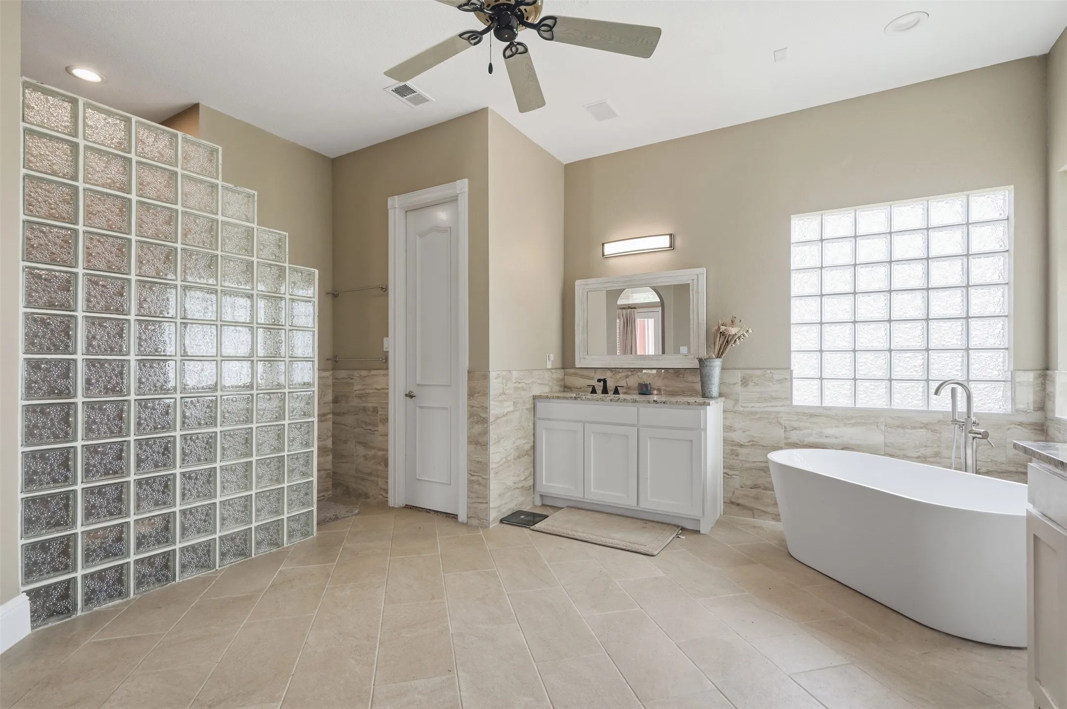 Spacious bath featuring a freestanding soaking tub, single vanity with light-toned countertop, floor-to-ceiling glass block shower enclosure, and large format tile flooring