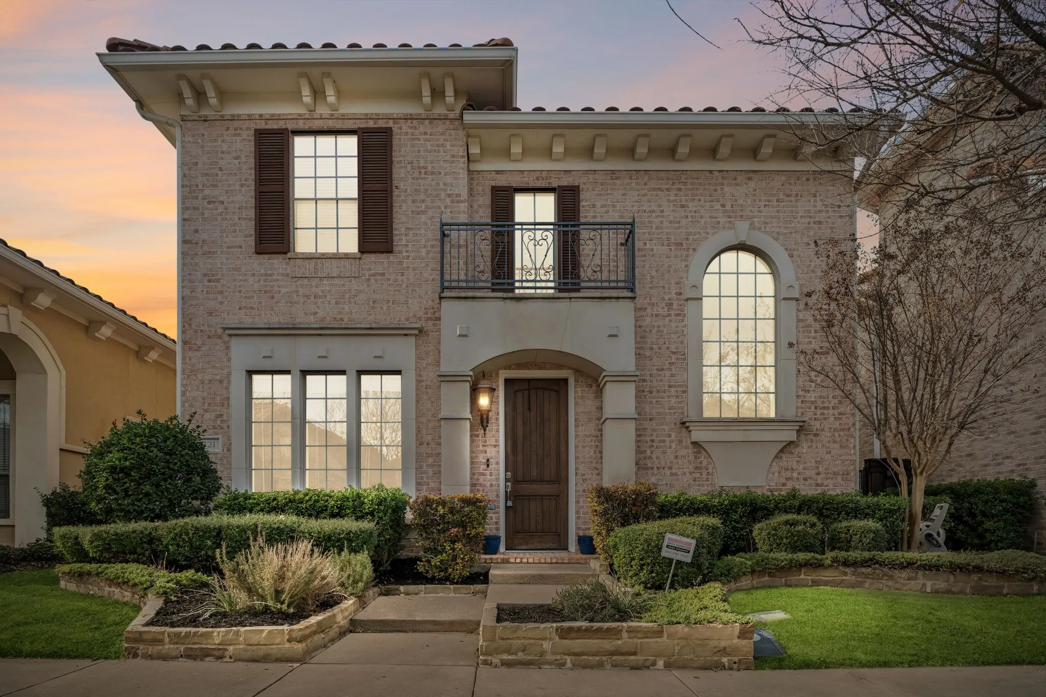 Brick exterior featuring a tile roof, decorative wrought iron balcony, arched window, and landscaped front yard with stone-edged planters