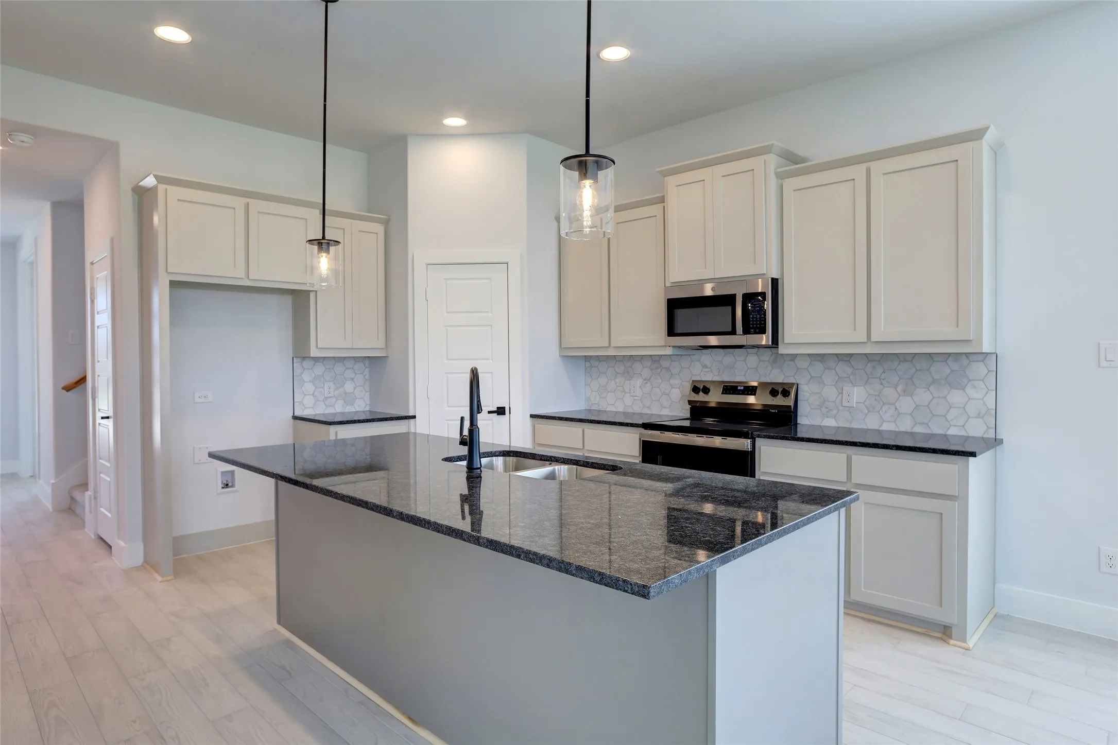 Kitchen featuring a central island with a dark countertop and undermount sink, light wood-finish flooring, light-toned cabinetry, stainless steel appliances, and a hexagonal tile backsplash