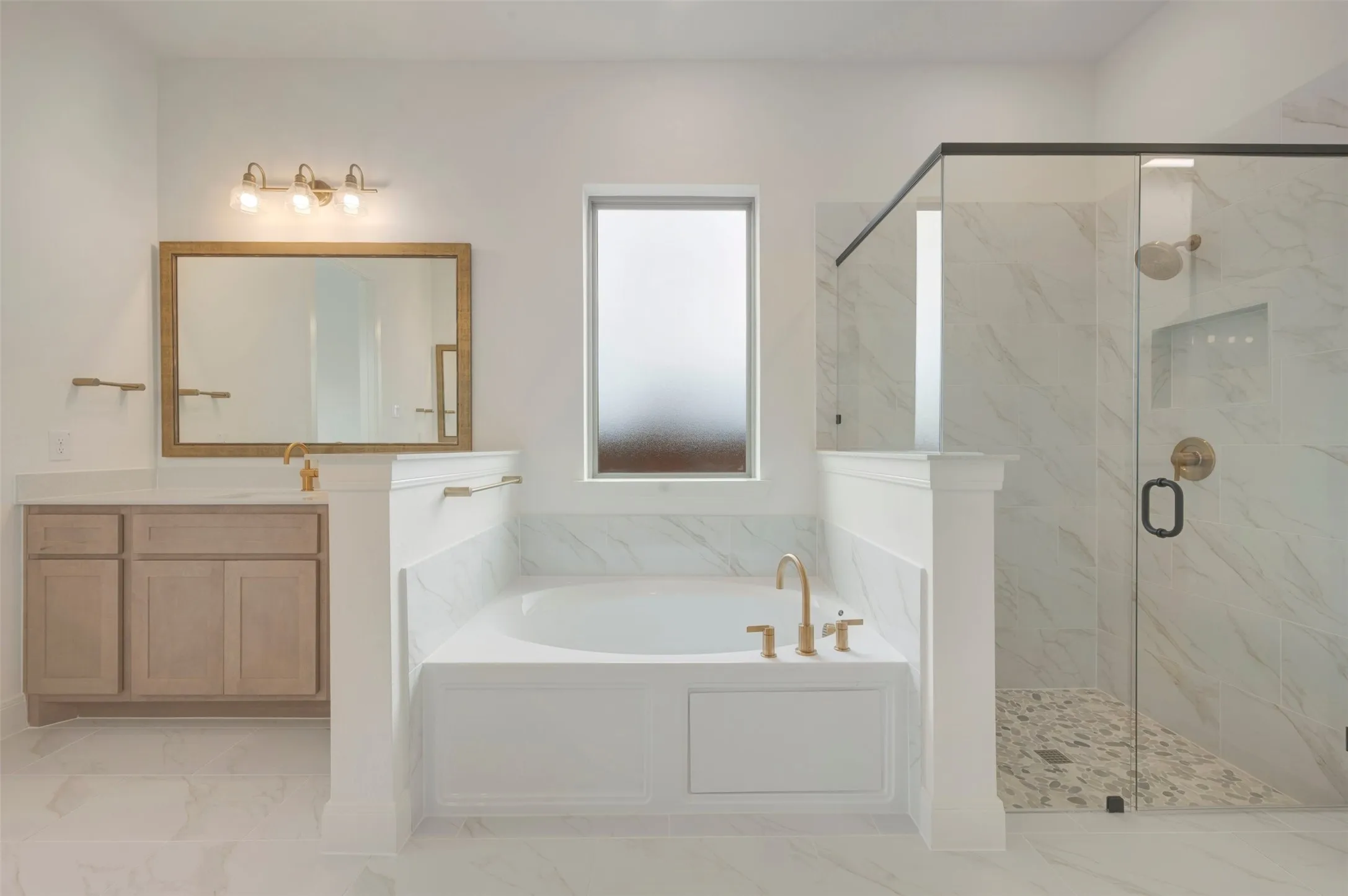 Contemporary bathroom featuring a freestanding tub, glass-enclosed shower, light wood-finish vanity, large framed mirror, and gold-tone fixtures