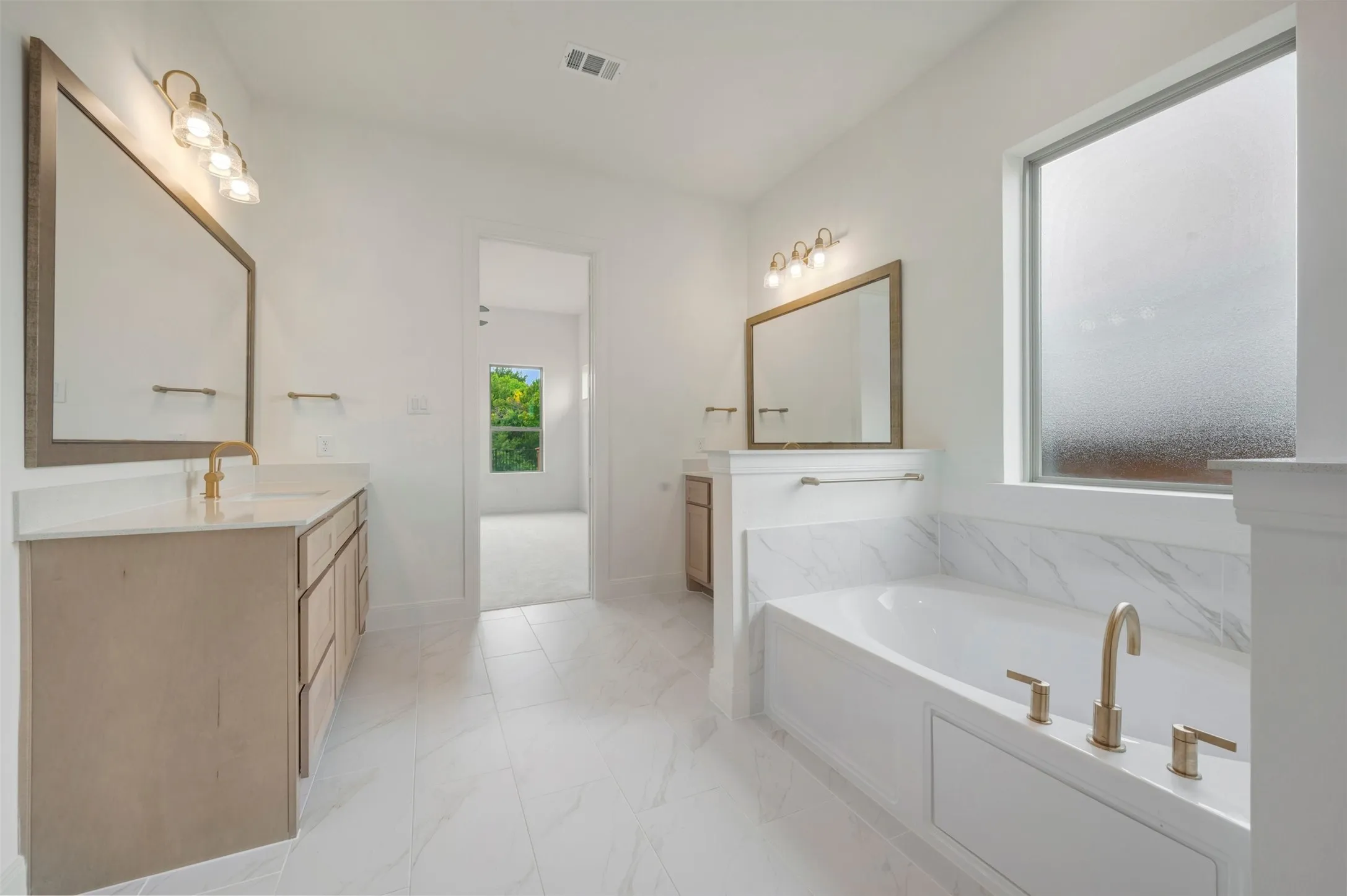 Bathroom featuring a built-in soaking tub with brass fixtures, dual vanities with light wood-finish cabinetry, a large framed mirror, and a tile shower surround