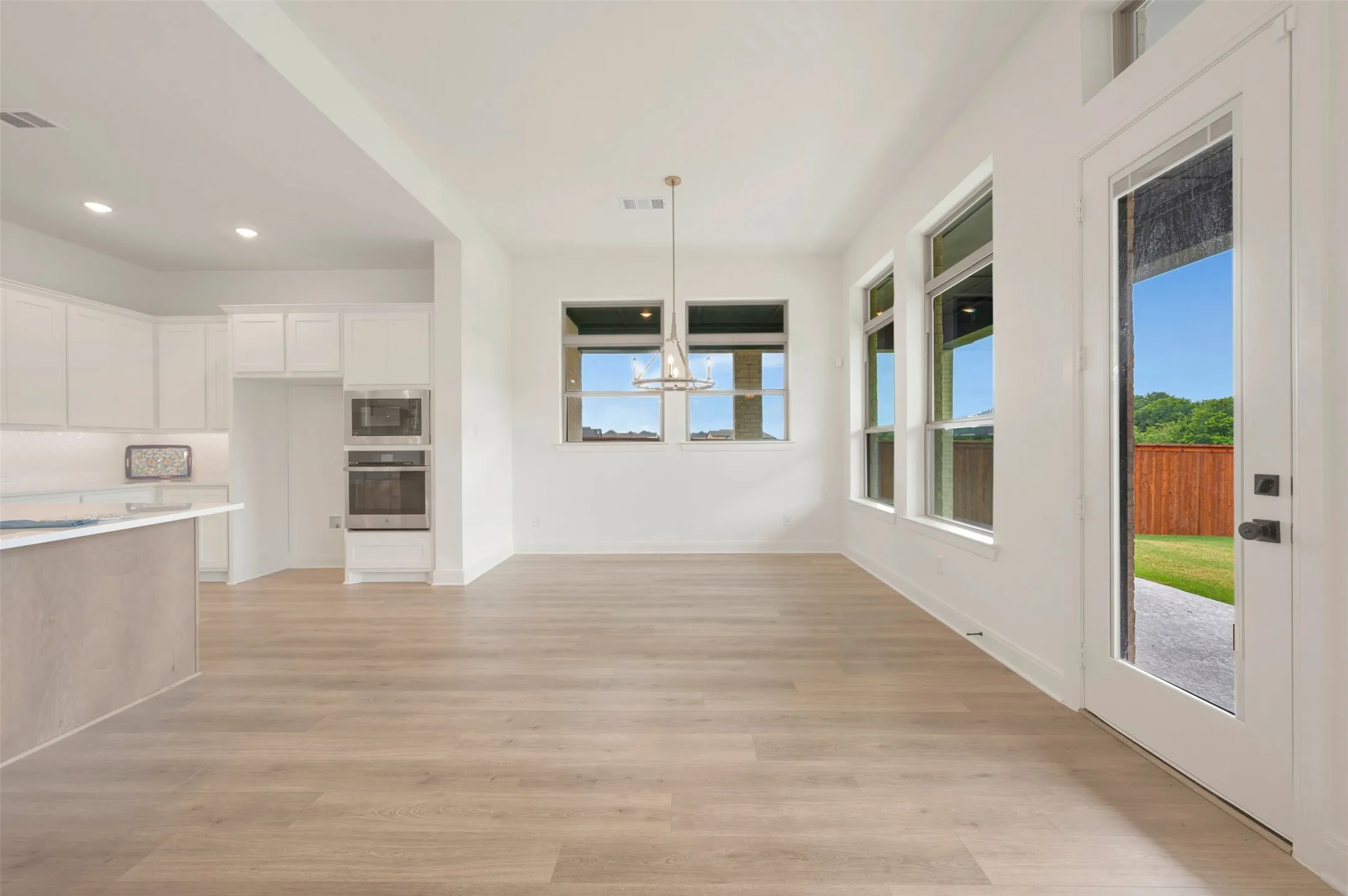 Open-concept kitchen and dining area featuring light wood-finish flooring, a contemporary chandelier, white cabinetry, integrated stainless steel double ovens, and large windows with a backyard entrance