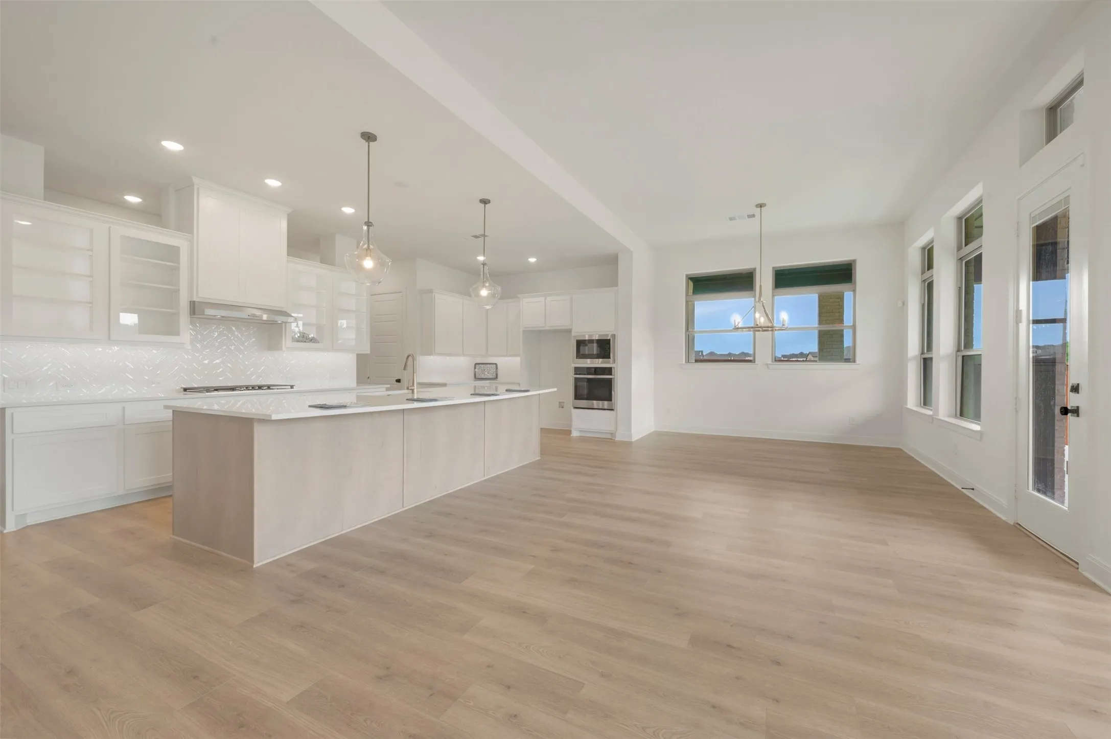 Open-concept living space featuring wood-finish flooring, a kitchen island with integrated sink, white cabinetry with glass-front uppers, a patterned tile backsplash, and built-in double ovens