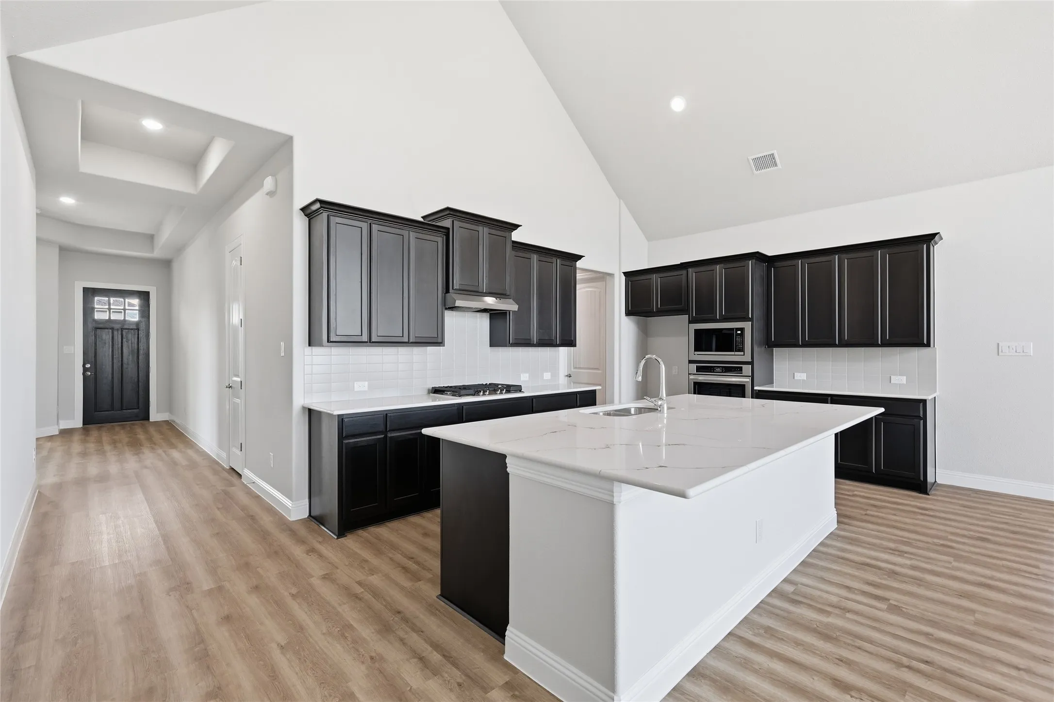 Kitchen with dark cabinets, backsplash, a center island with sink, light stone counters, and vaulted ceiling