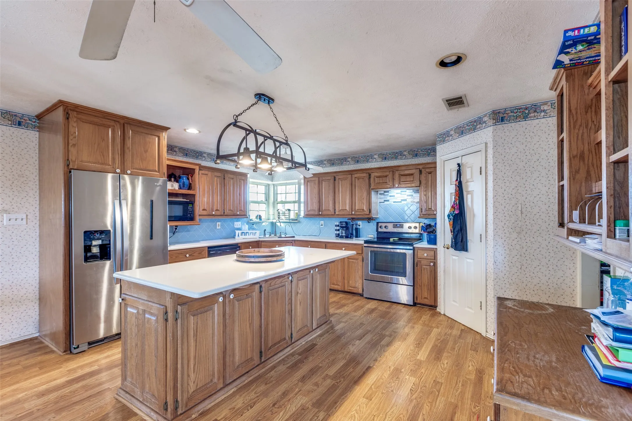 Kitchen with wallpapered walls, wood finish cabinets, stainless steel appliances, light countertops, and open shelves