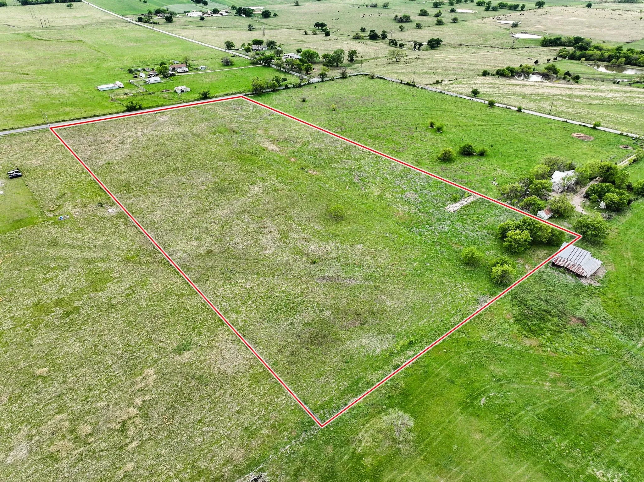 Expansive green acreage featuring a clear perimeter, a rustic outbuilding with a corrugated metal roof, mature trees, and surrounding open fields