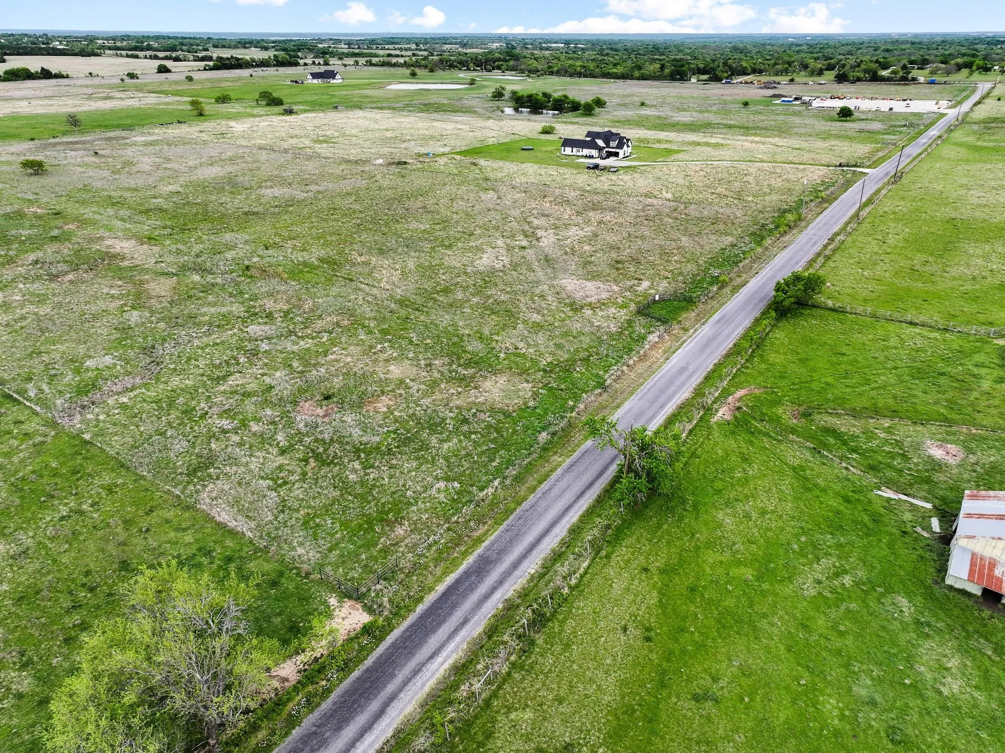 Expansive acreage featuring a paved road frontage, scattered mature trees, and a distant single-story dwelling with a dark roof