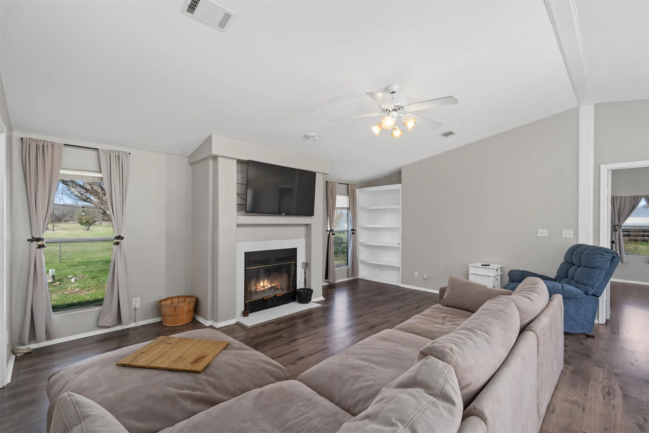 Living area featuring dark wood-style floors, healthy amount of natural light, vaulted ceiling, a ceiling fan, and a glass covered fireplace
