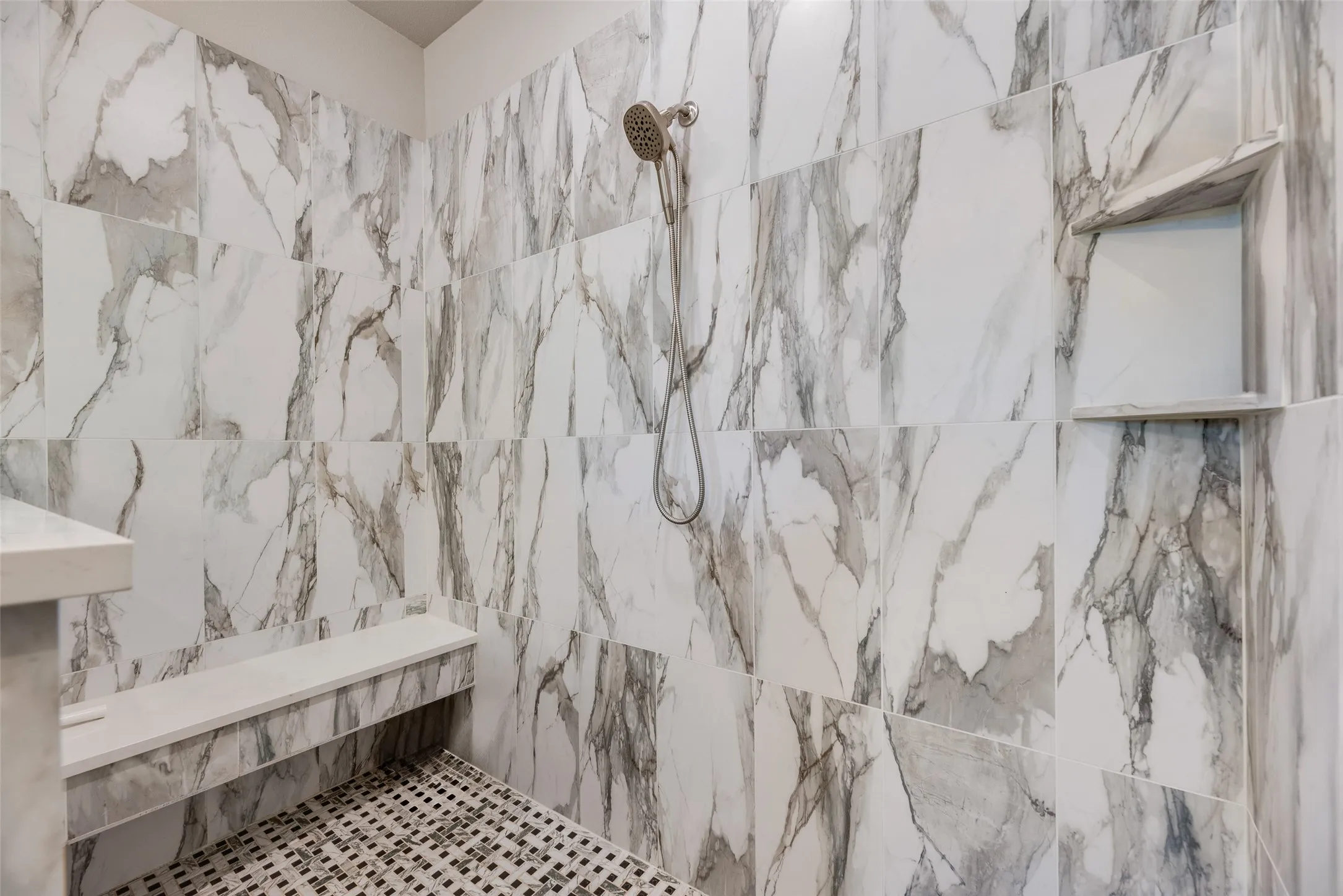 Shower featuring large-format tile walls with prominent veining, a built-in bench, a recessed niche, a handheld showerhead, and a mosaic tile floor