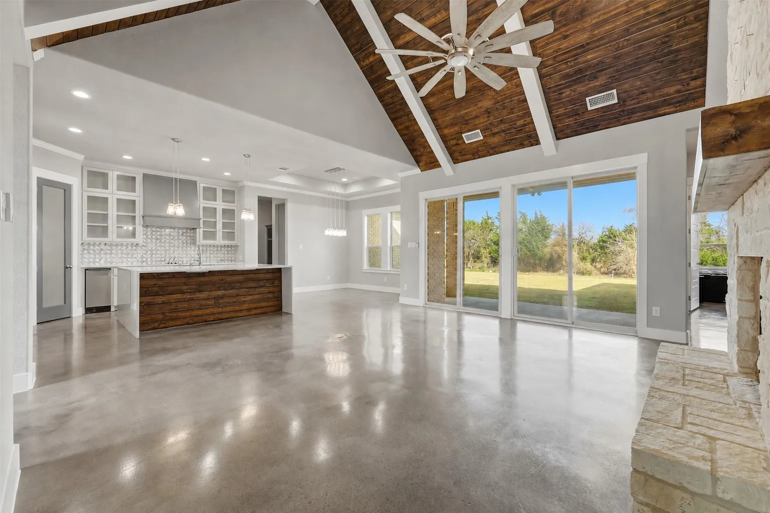Vaulted wood-finish ceiling with exposed beams, polished concrete flooring, extensive sliding glass doors, a stone fireplace, and a kitchen island with a wood-finish base