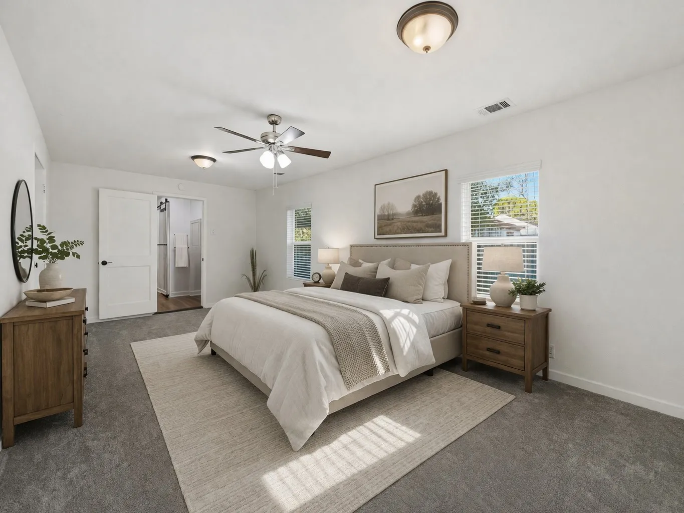Spacious bedroom featuring light gray carpeting, neutral wall colors, dual windows with blinds, a ceiling fan with integrated lighting, and a barn door entry to an adjoining bathroom - Virtually staged