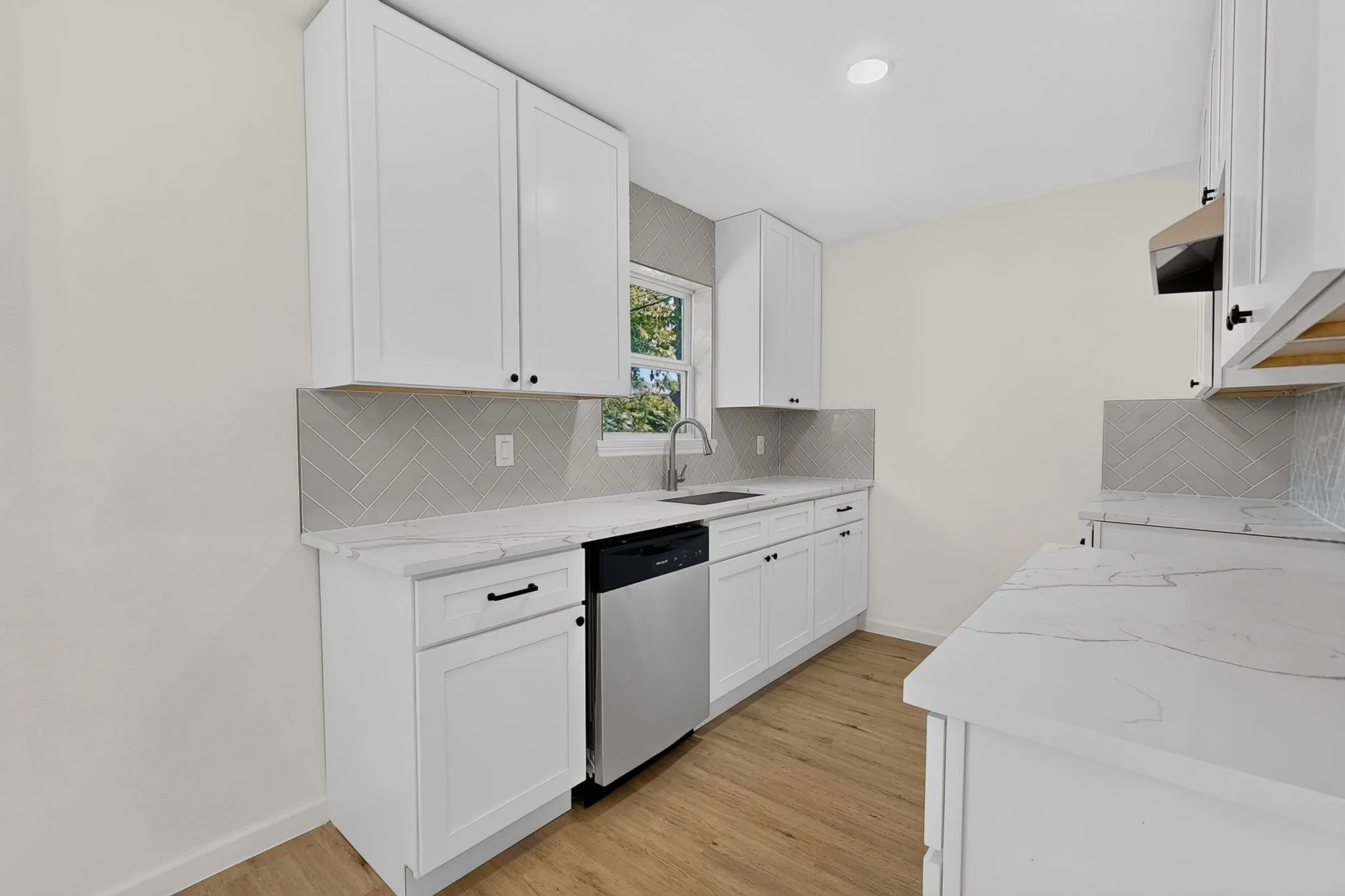Contemporary kitchen featuring white cabinetry with black hardware, white countertops, a gray herringbone tile backsplash, and wood-finish flooring