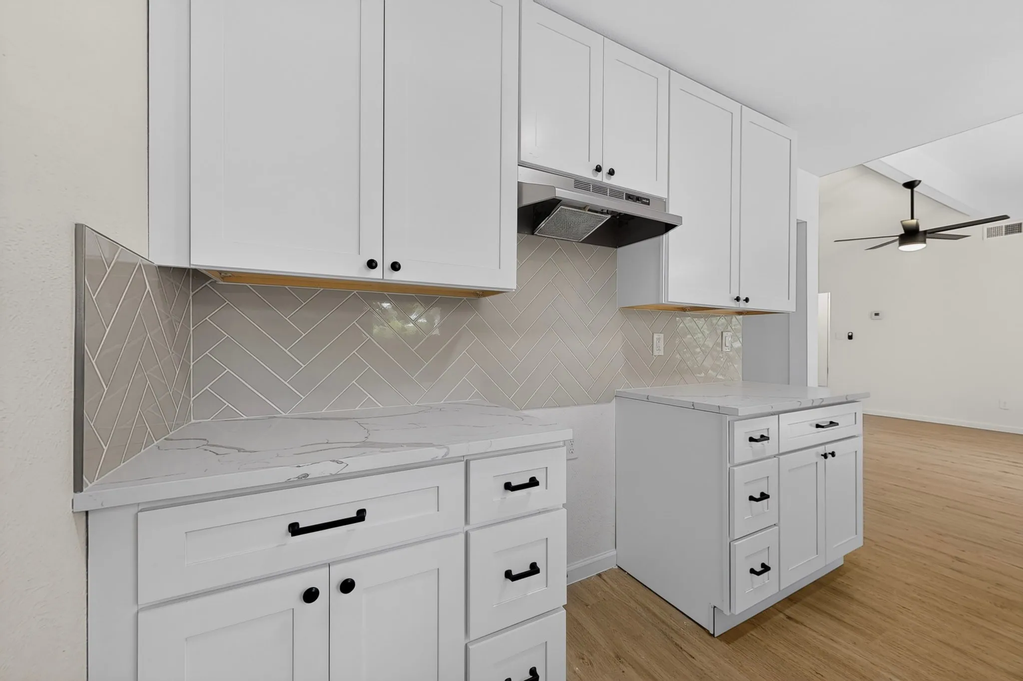 White shaker cabinetry with dark hardware, stone-finish countertops, herringbone tile backsplash, under-cabinet lighting, and wood-finish flooring