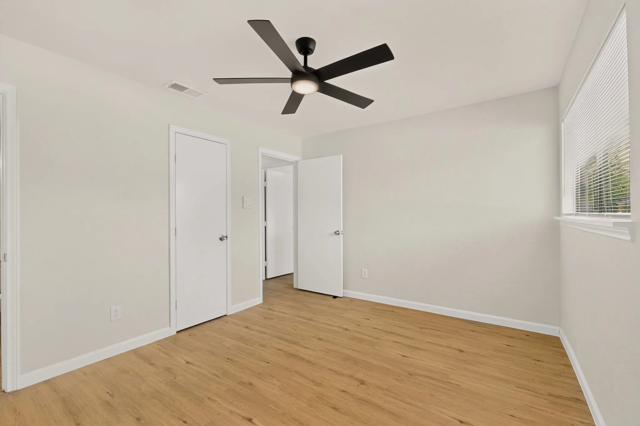 Spacious room featuring wood-finish flooring, light neutral wall tones, and a modern ceiling fan with integrated lighting