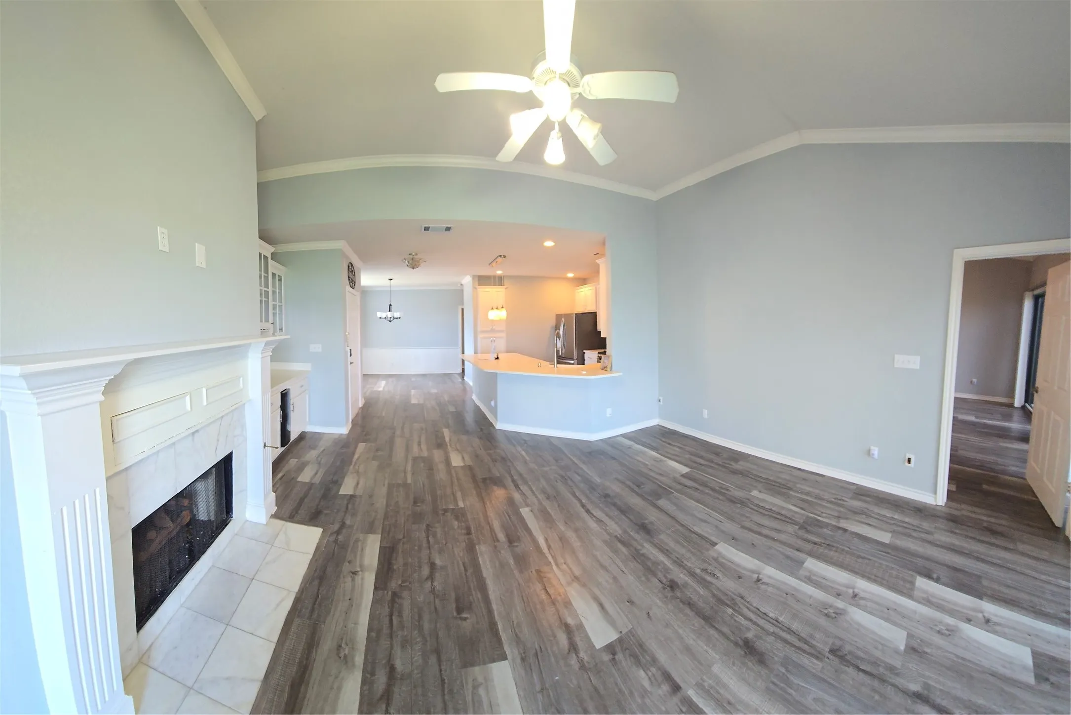 Spacious living area featuring wood-finish flooring, a white mantel fireplace with a tile hearth, vaulted ceilings with crown molding, and an integrated ceiling fan