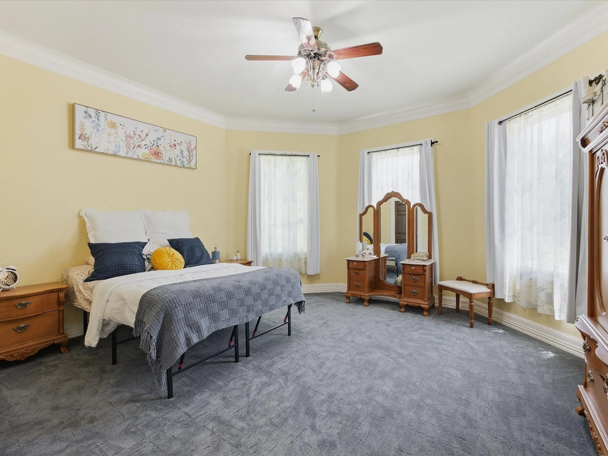 Spacious room featuring gray patterned carpeting, multiple windows with white sheer curtains, substantial crown molding, and a ceiling fan with integrated lighting