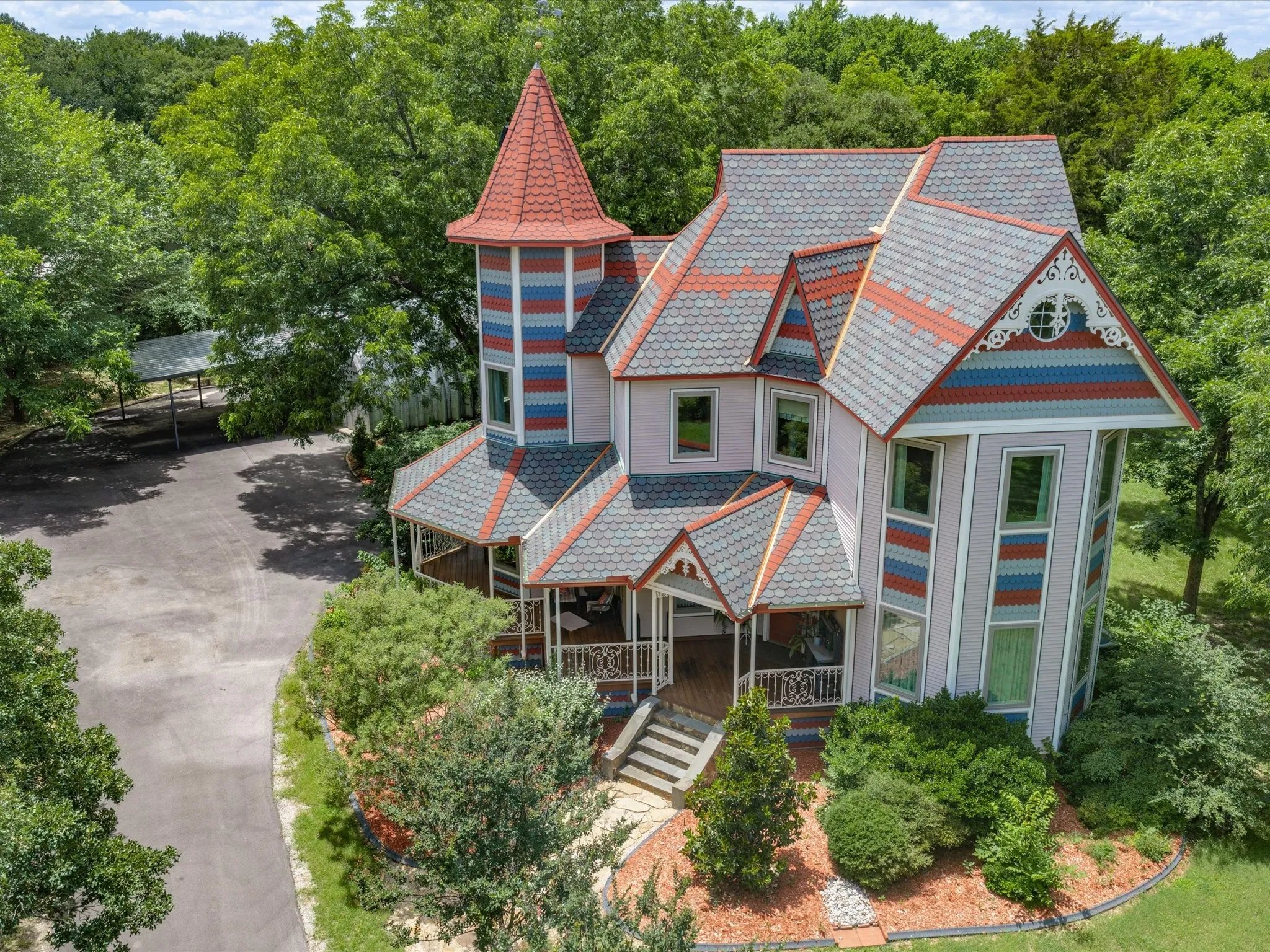 Victorian-style exterior featuring a prominent turret with a red tile roof, multi-color siding accents, multiple covered porches, ornate gables, and a paved driveway leading to a carport