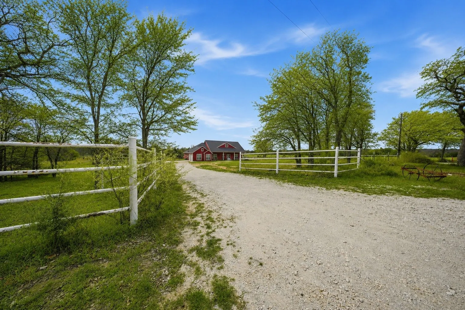 Gravel driveway leading to a red-sided residence with a dark roof, framed by mature trees and white fencing