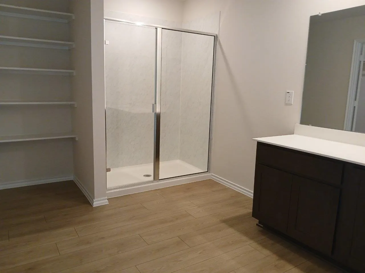 Bathroom featuring a glass-enclosed shower, wood-finish flooring, built-in shelving, a dark wood-finish vanity with a light-toned countertop, and a wall-mounted mirror