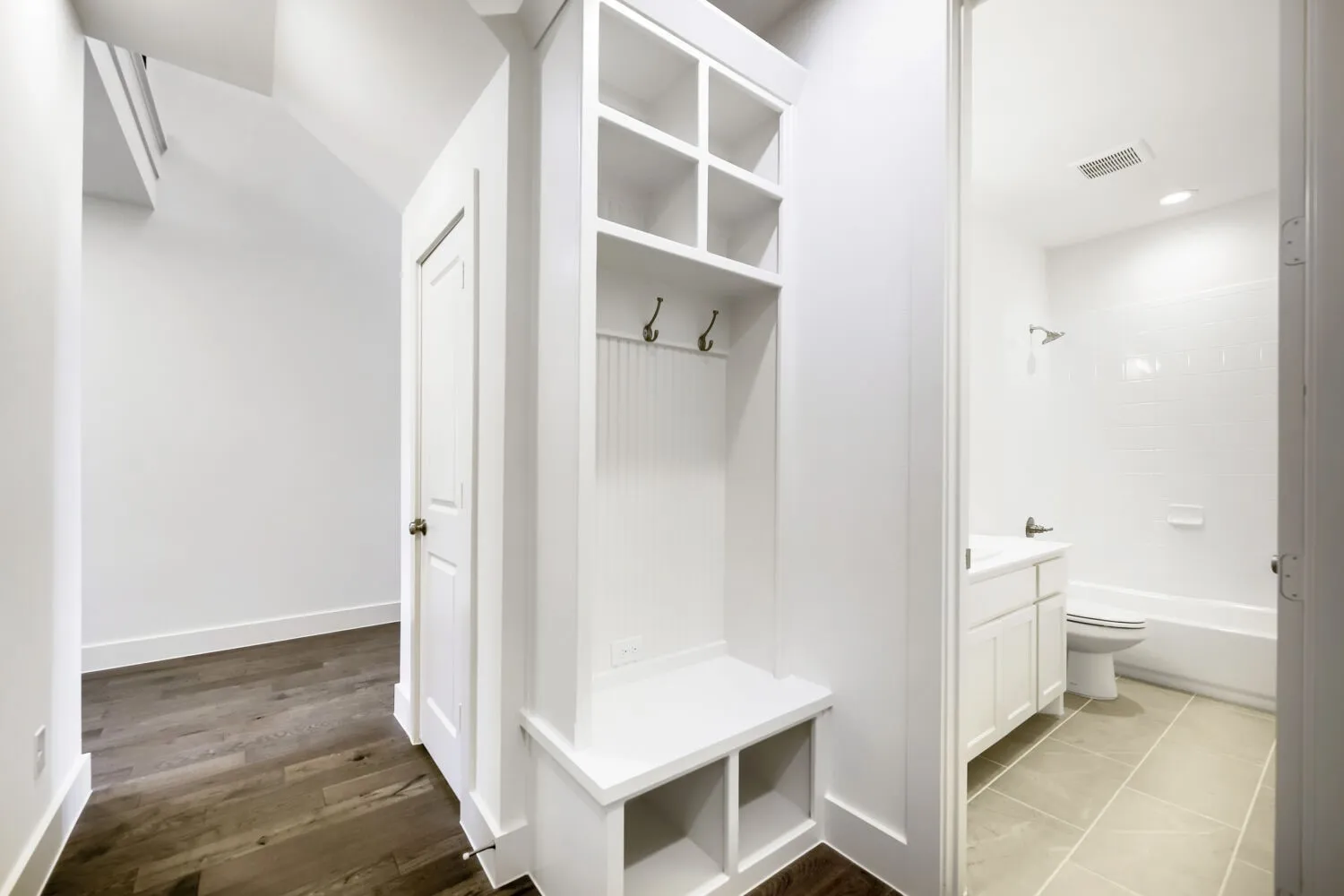Hallway featuring wood-finish flooring, a built-in storage bench with coat hooks and cubbies, a paneled door, and a full bathroom with a tiled floor, white vanity, and tub with subway tile surround
