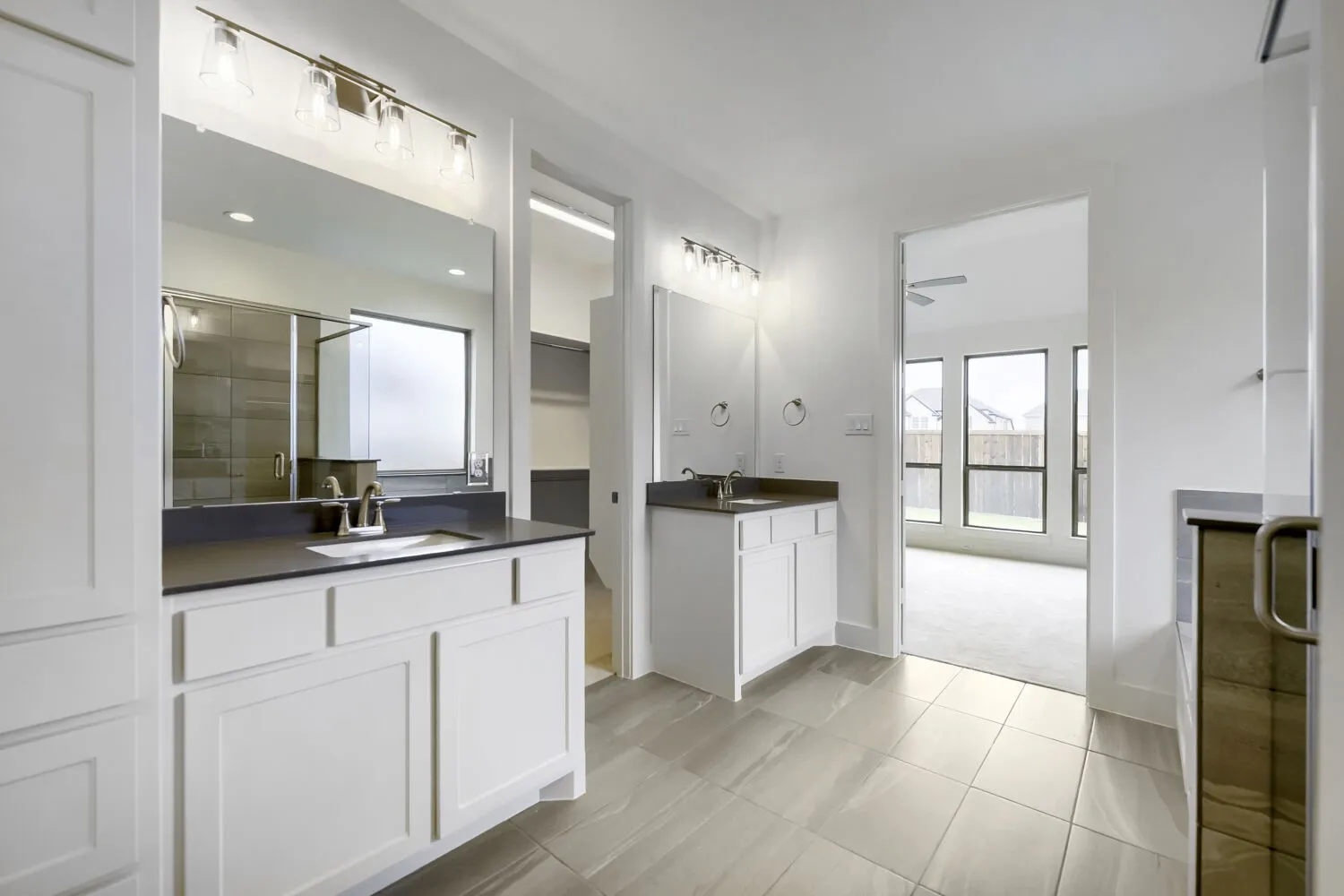 Bathroom featuring light-toned tile flooring, dual vanities with dark countertops, and white cabinetry