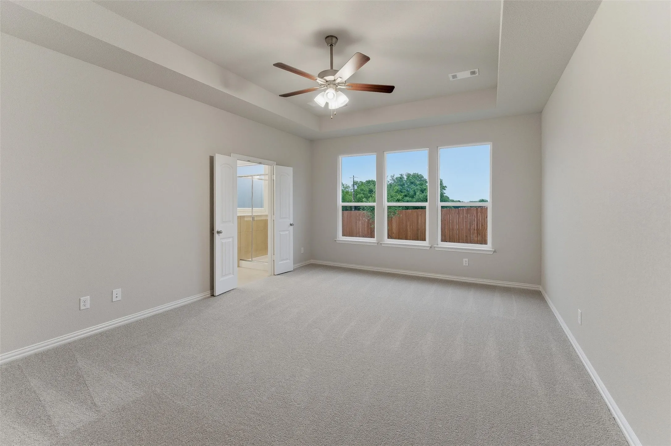 Spacious room featuring light-toned carpeting, a tray ceiling with a built-in ceiling fan, and three large windows providing abundant natural light