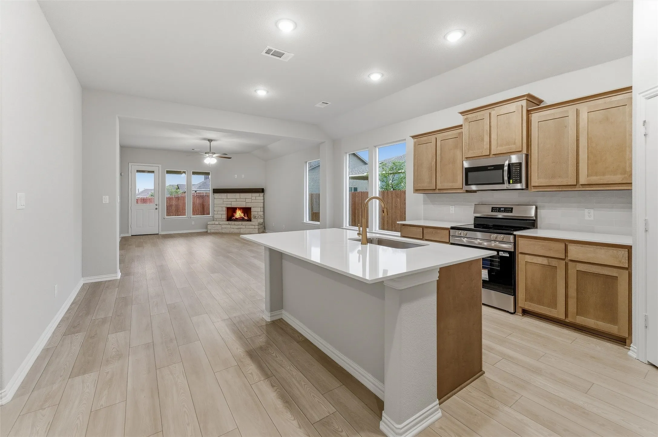 Spacious open-concept kitchen and living area featuring wood-finish flooring, a contemporary kitchen island with a single-basin sink, light wood cabinetry, stainless steel appliances, and a stone fireplace