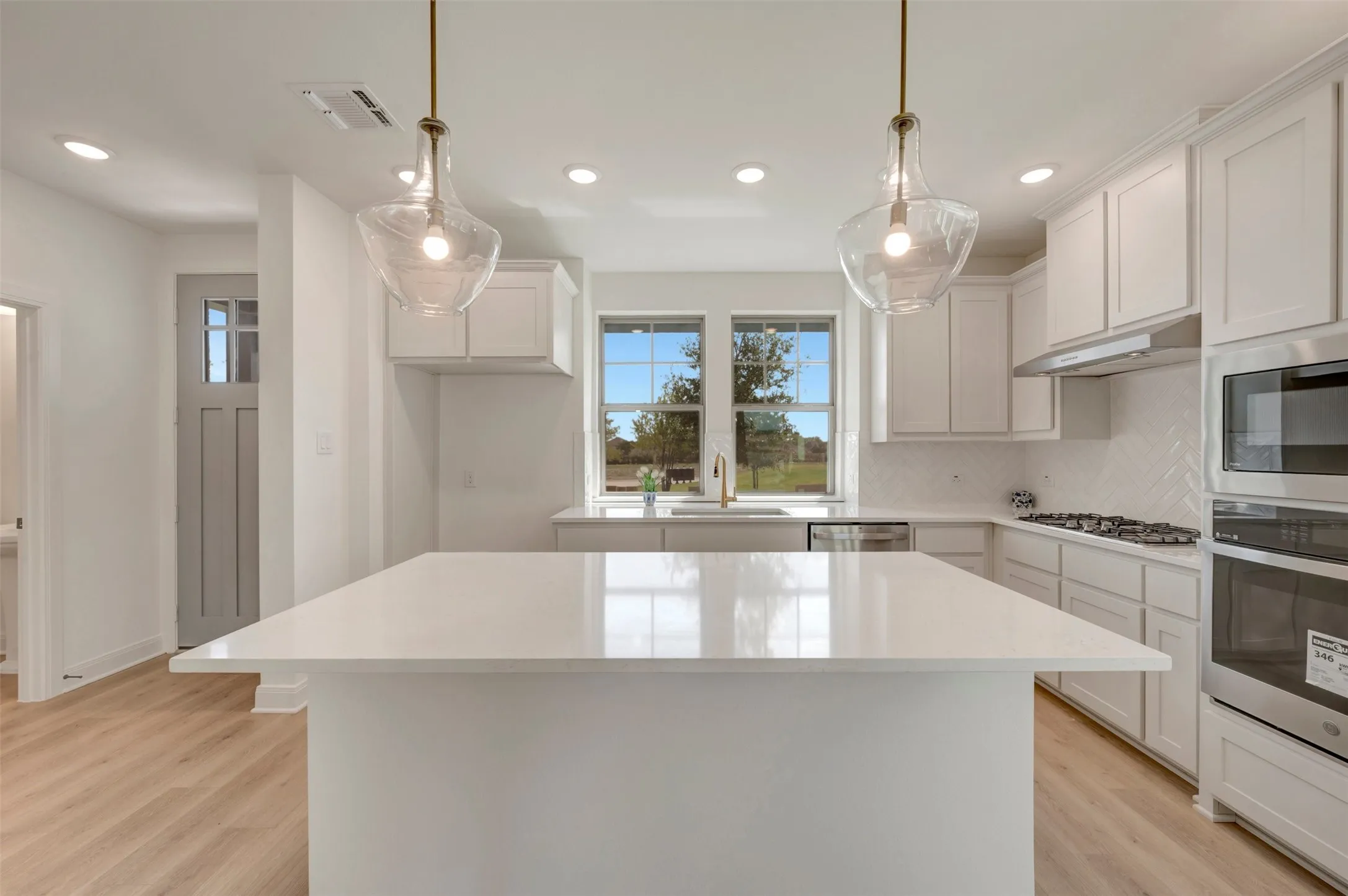Kitchen featuring a spacious island with a light-toned countertop, white cabinetry, integrated stainless steel appliances, herringbone-patterned tile backsplash, and recessed lighting