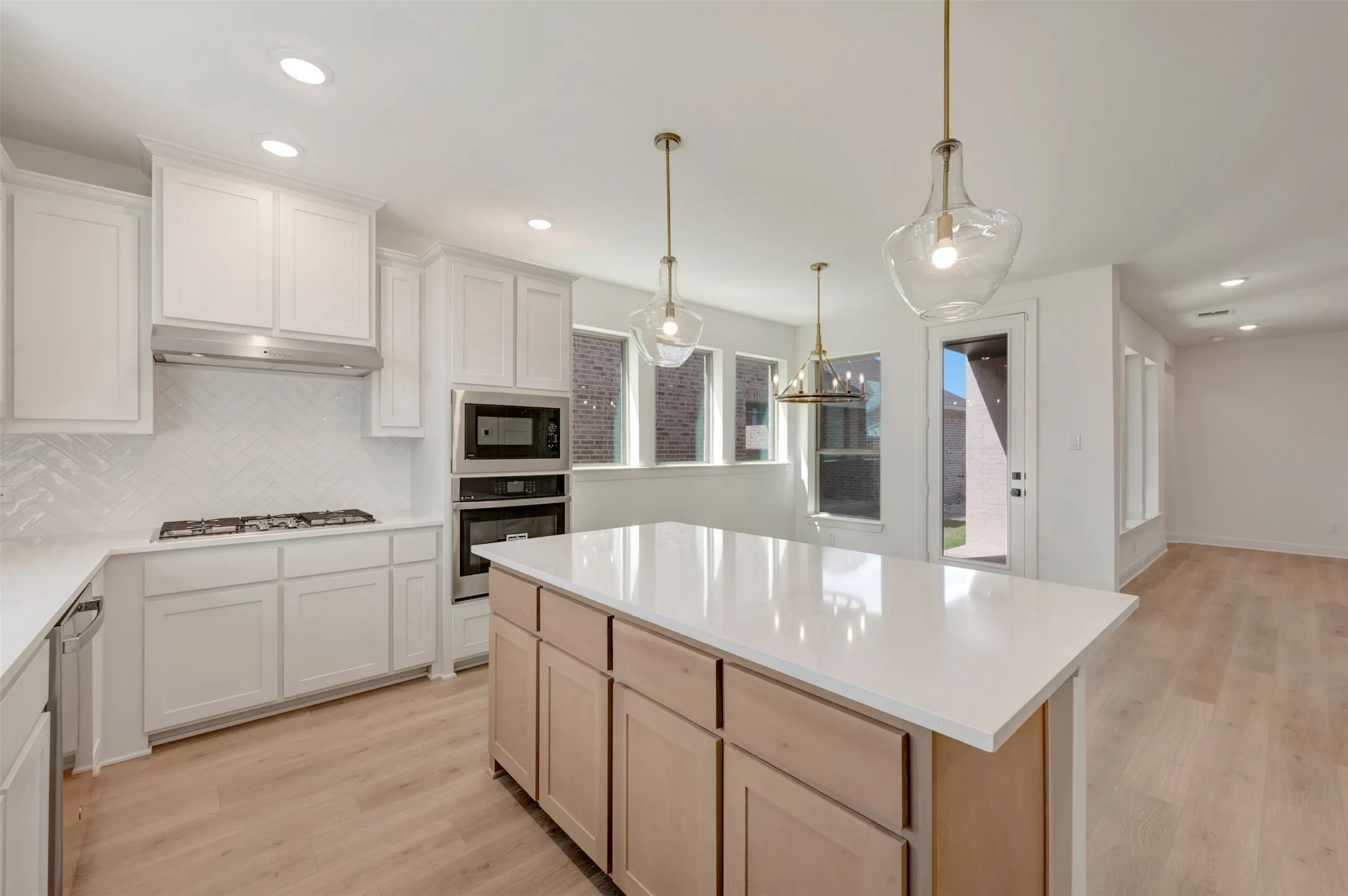 Bright kitchen featuring a substantial island with a light-toned countertop, white cabinetry with crown molding, stainless steel built-in appliances, a herringbone tile backsplash, and wood-finish flooring